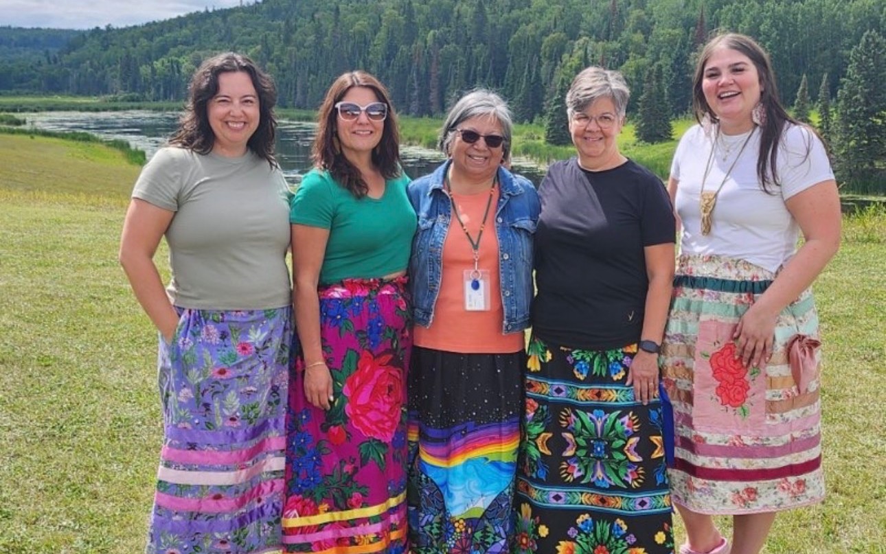 WNMB Research Team Members Céline Wick and four other women, wearing t-shirts and Métis ribbon skirts, stand outdoors; trees and a river are visible in the background