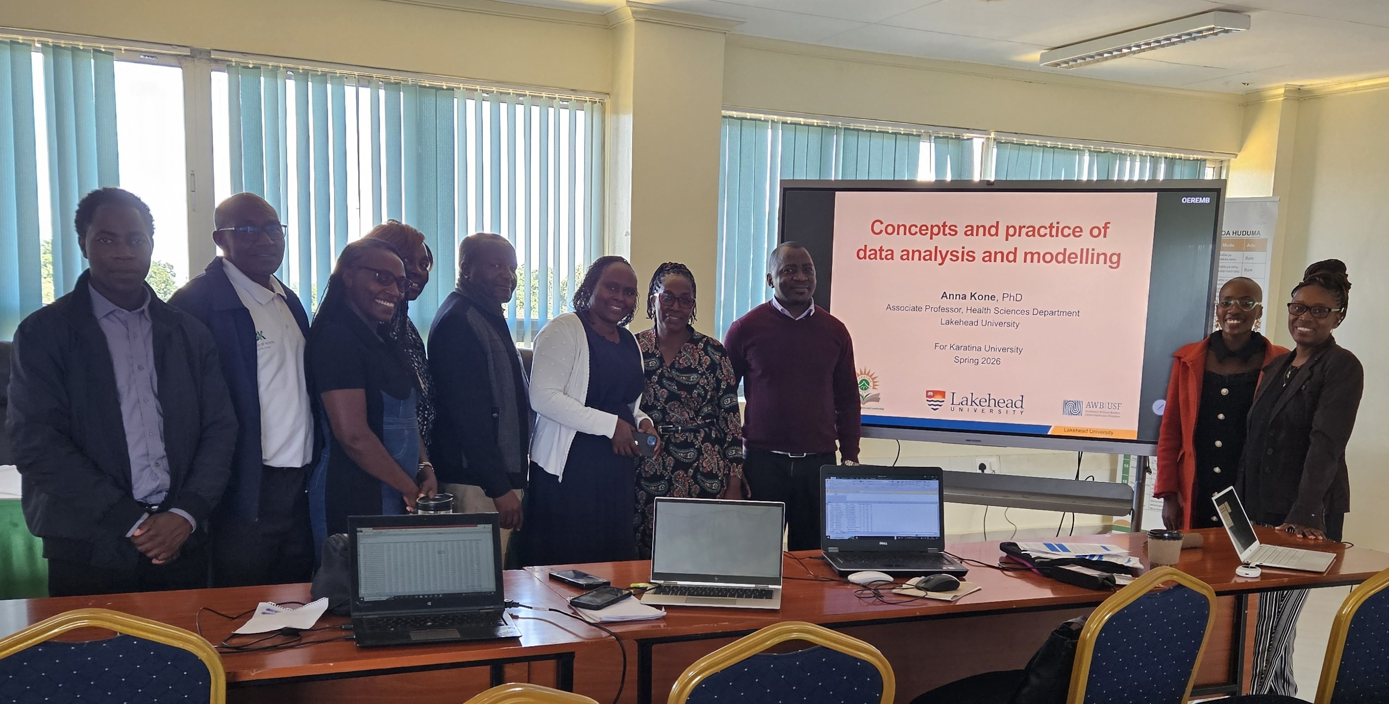 A group of people stand behind a table in a conference room with a video screen in the background saying "Concepts and practice of data analysis and modelling"