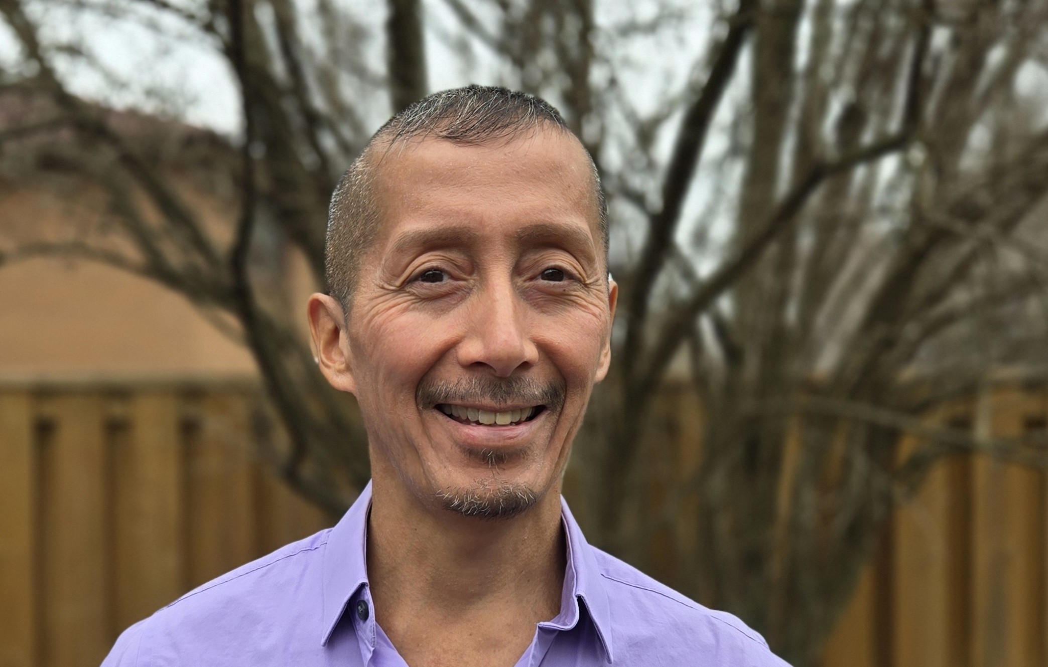 A head-and-shoulders photo of Dr. Gerardo Reyes, wearing a purple shirt, standing in a yard with a tree and a wooden fence visible in the background