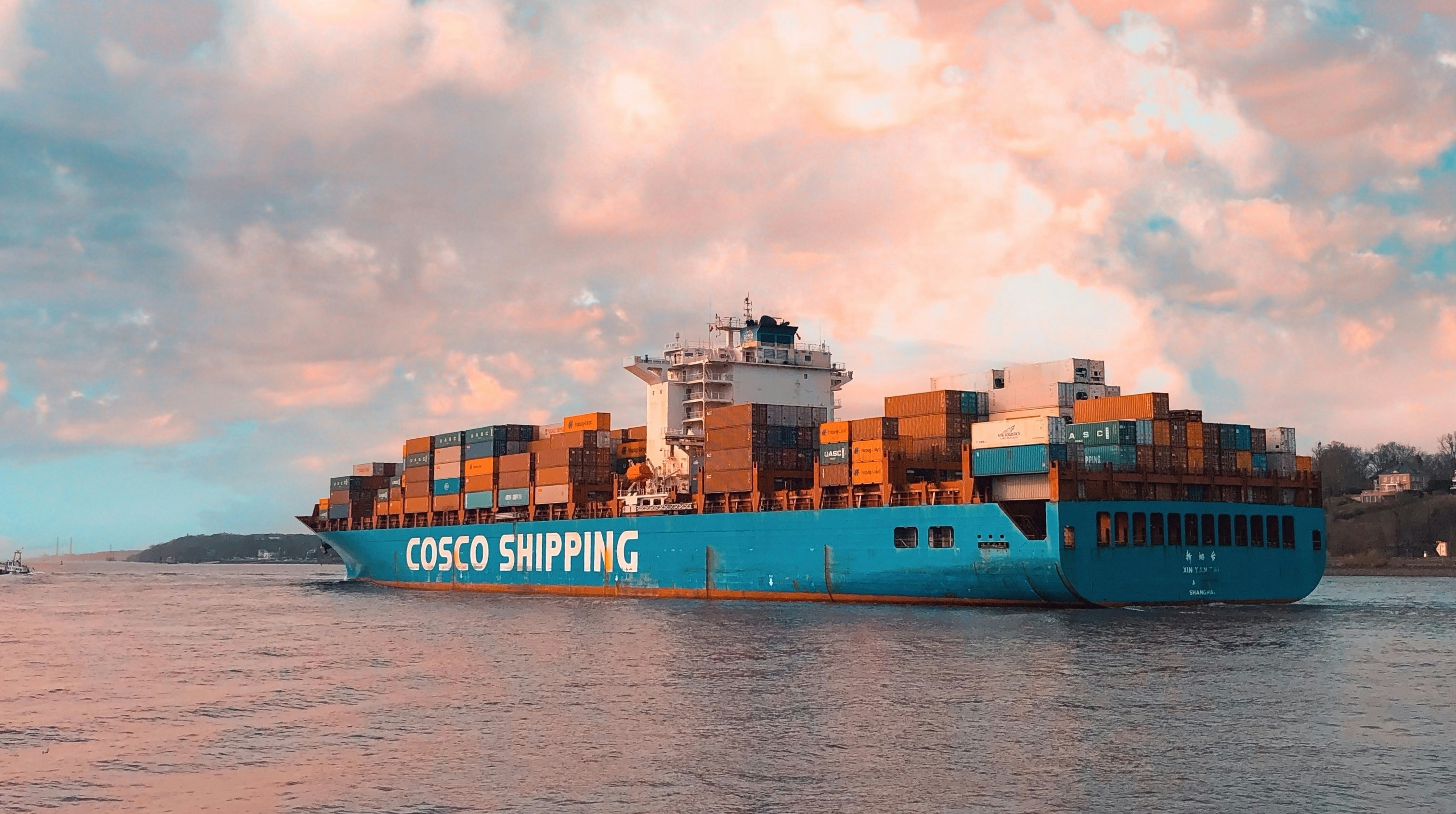 A large blue cargo ship stacked with many rectangular cargo containers sails near a coastline with trees and a house