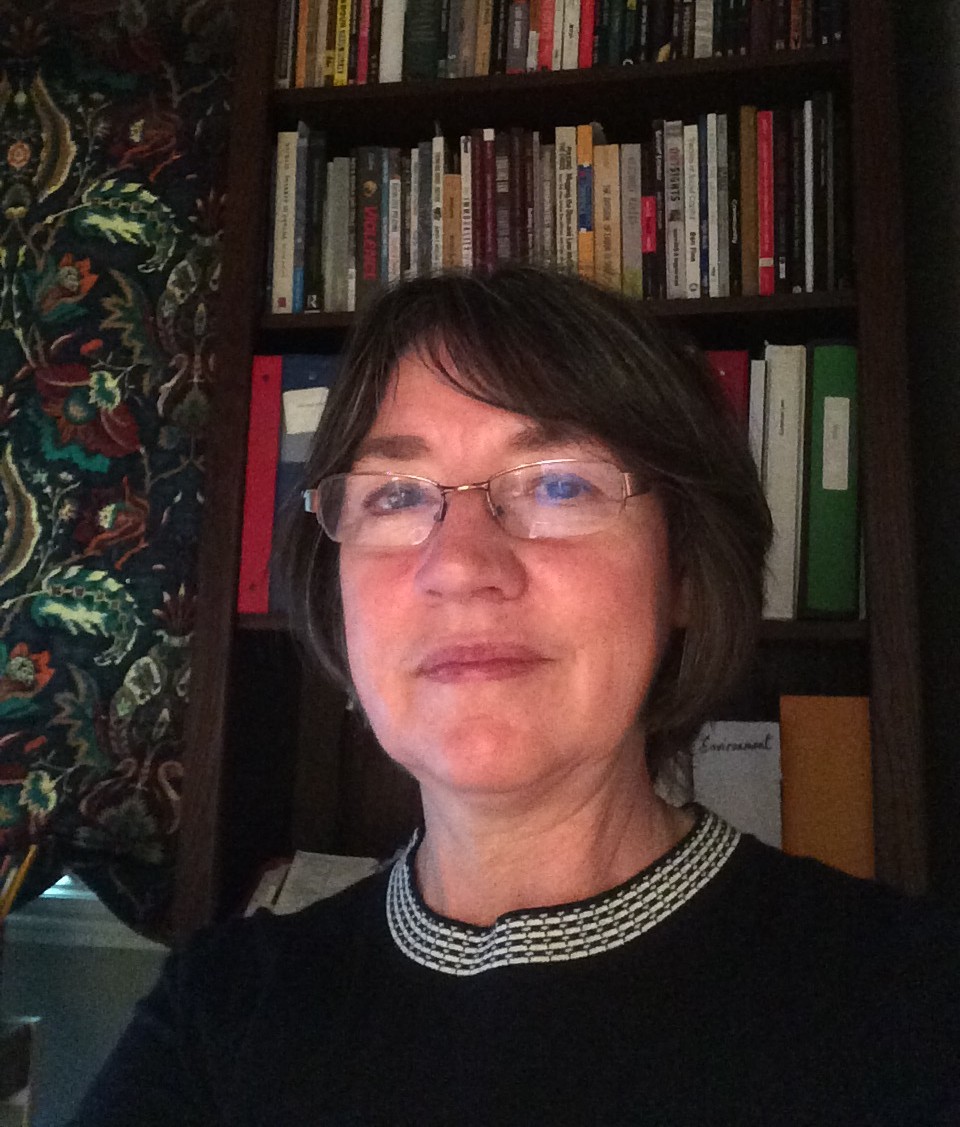Dr. Jennifer Jarman, wearing glasses and a black top with a white-and-black collar stands in front of a bookcase