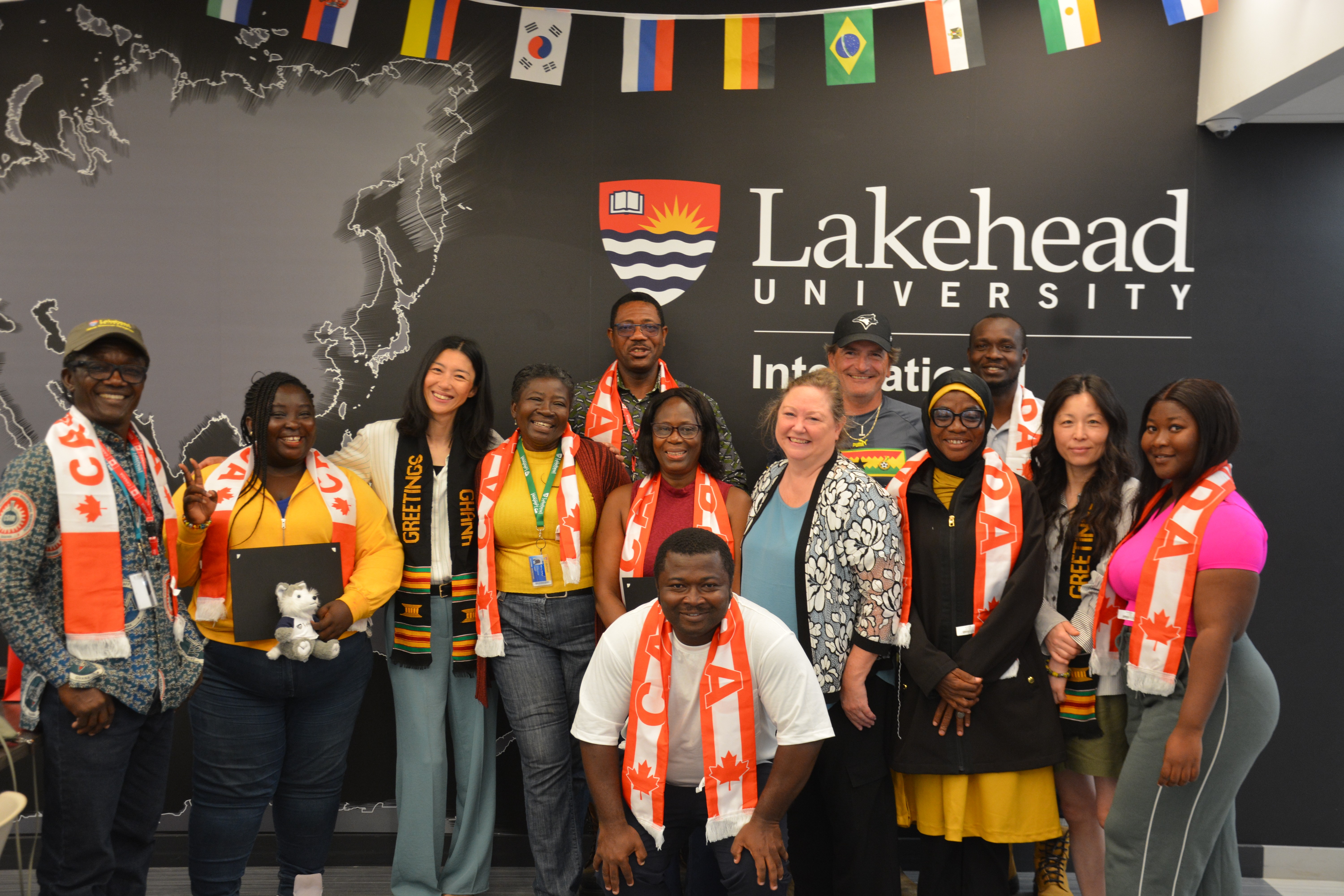 A group of smiling people wearing scarves saying "Canada" stand in front of a wall emblazoned with the words "Lakehead University International"