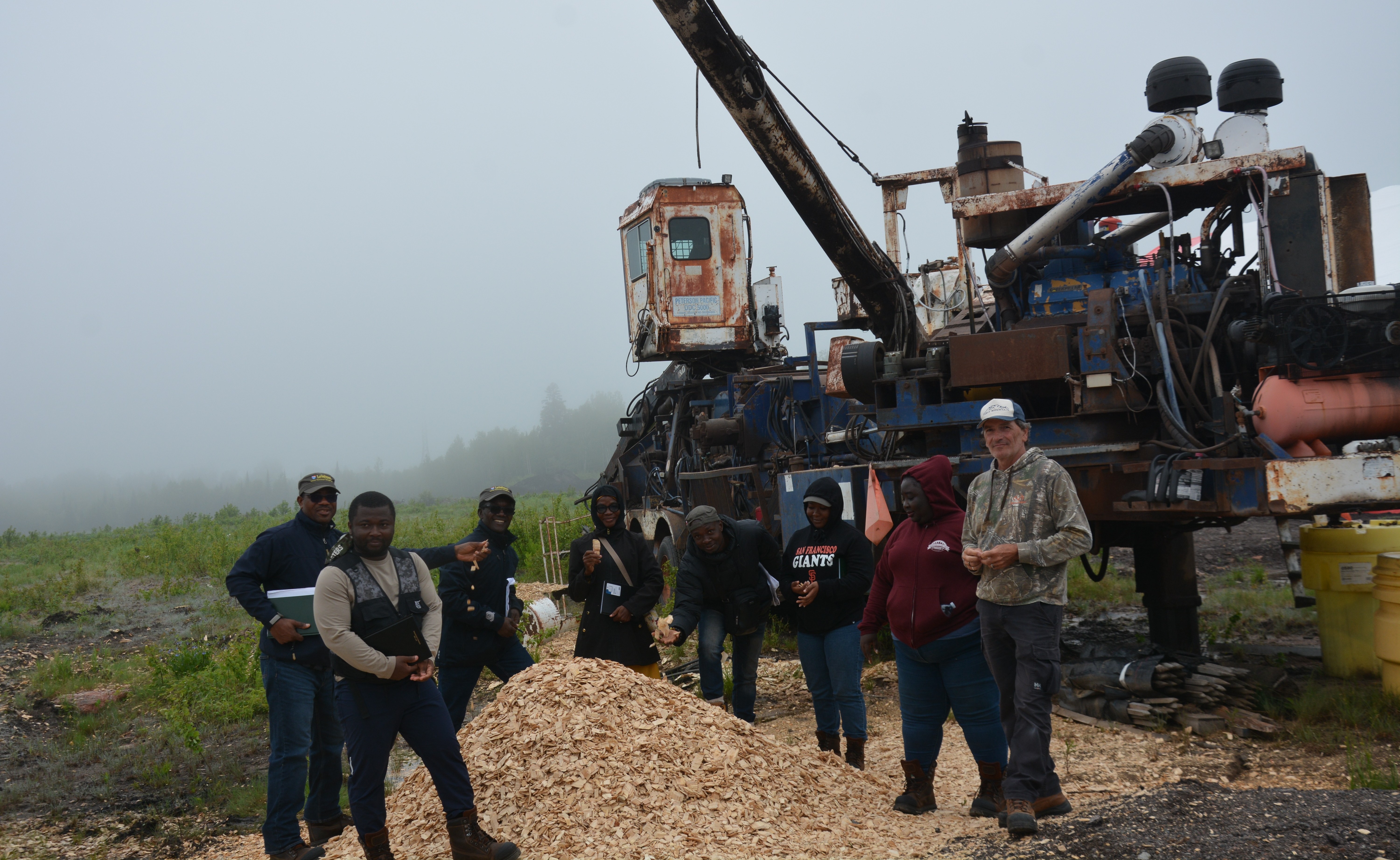 A group of casually dressed people stand outside on a foggy day beside a pile of wood chips and a large piece of forestry equipment