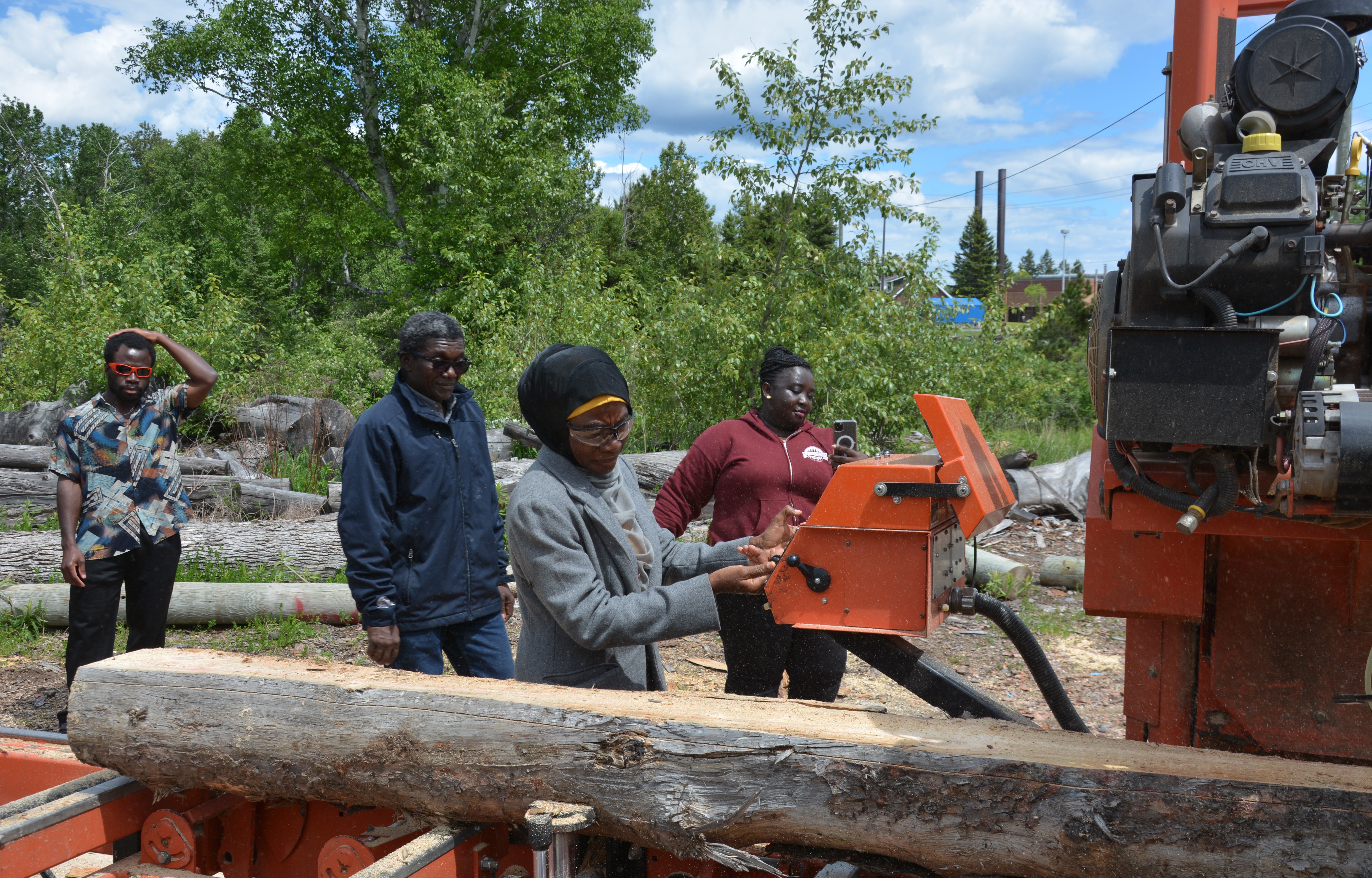 A woman operates a portable milll while three other people look on; logs and a forested area are visible in the background