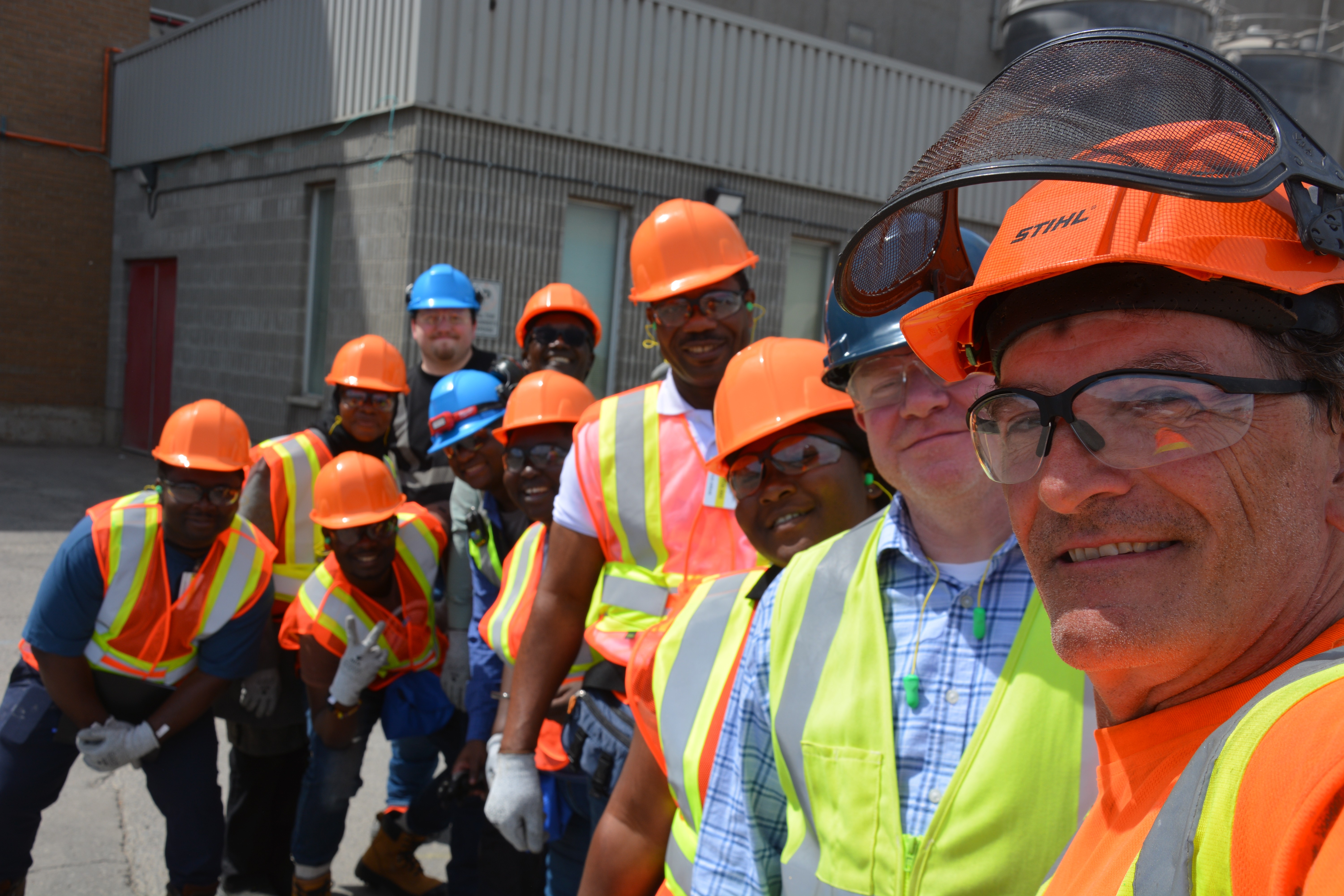 A group of people wearing safety vests, hard hats, and safety glasses stand in front of an industrial building