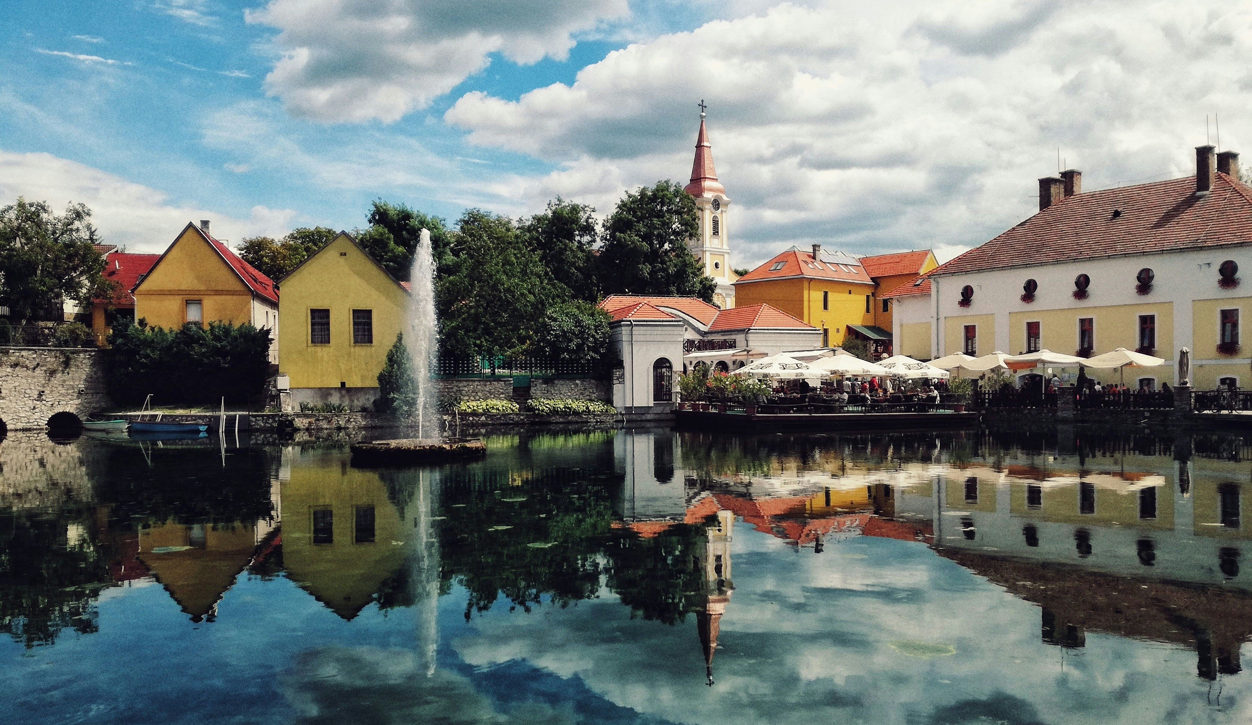 Historic stone and stucco buildings, including a church, surround a large pond on a sunny day; people sit under canopied tables near the water's edge