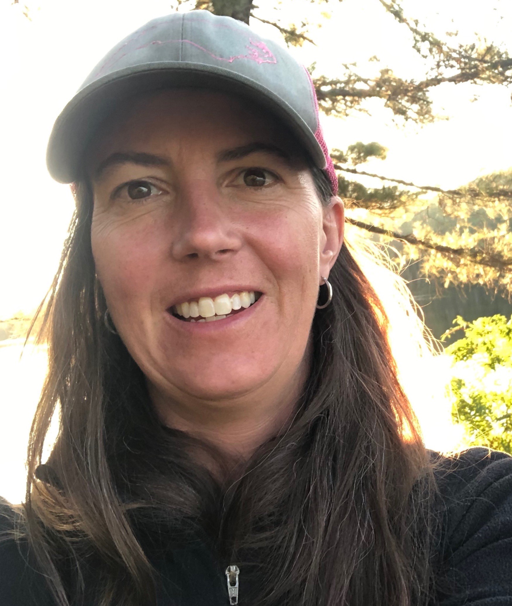 A head-and-shoulders photo of Linsday Sargent outside wearing a grey fleece zip-up, baseball cap, and hoop earring; a tree and a lake are visible in the background