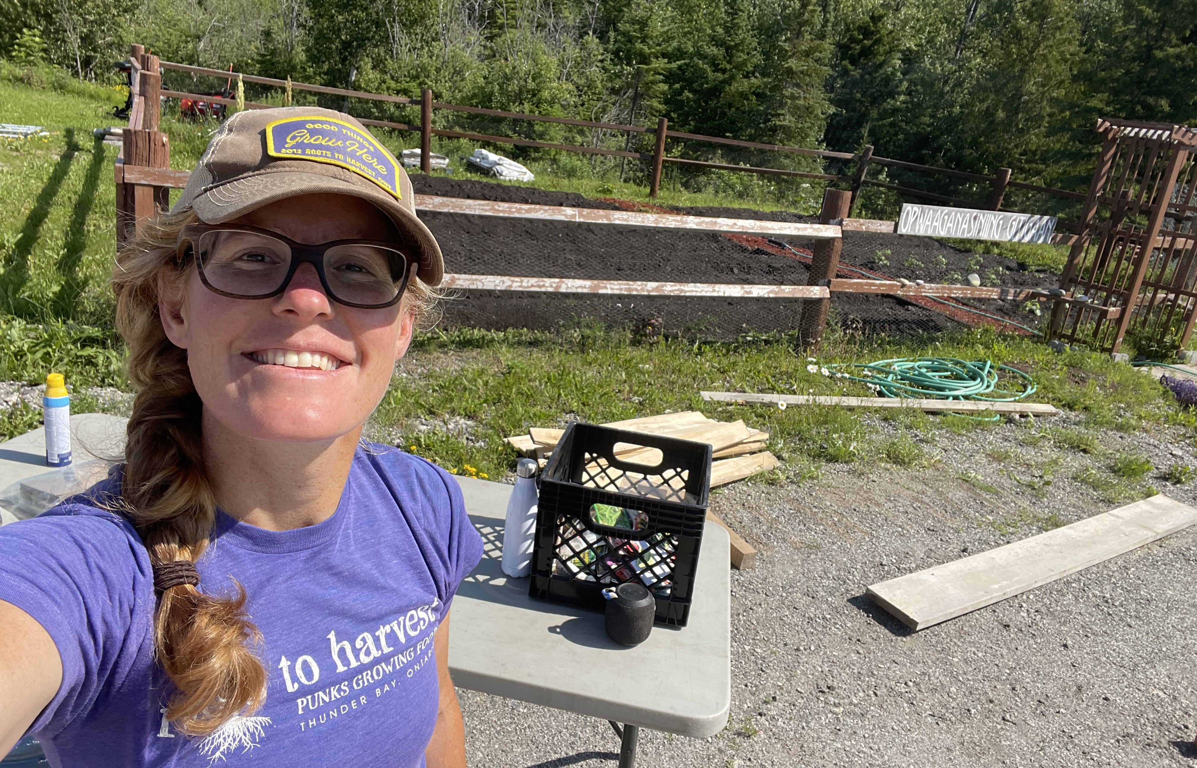 Erin Beagle, wearing a baseball cap and glasses, stands beside a fenced-in garden