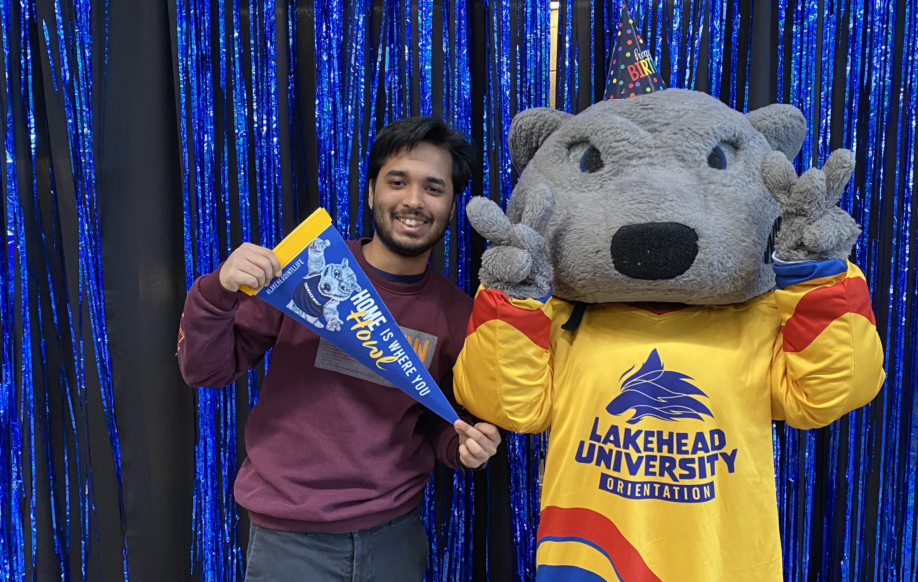 Faiz Mahmud holds a pennant saying Home is Where You Howl and stands next to the Wolfie mascot who is holding both hands up in a peace sign