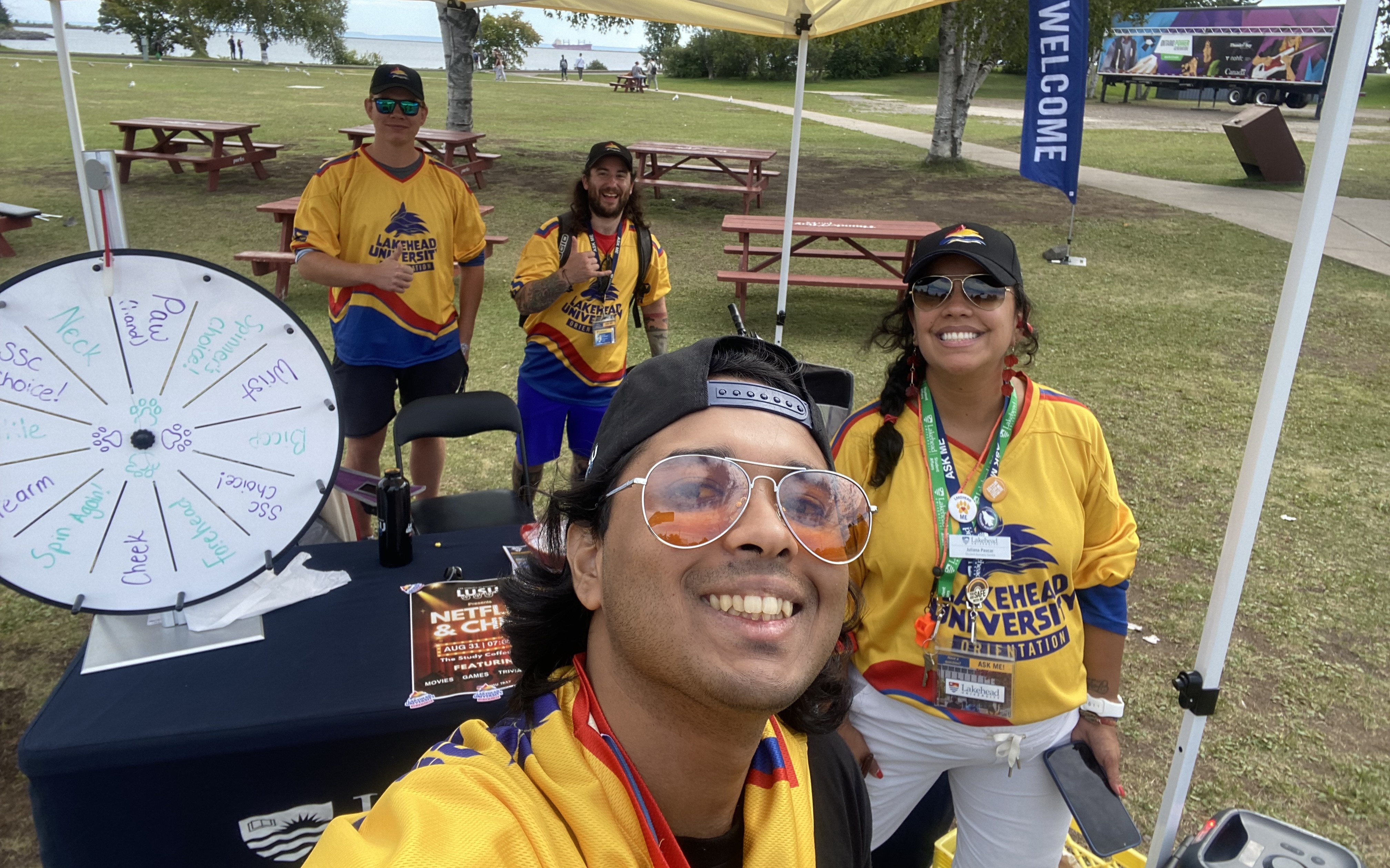 Faiz Mahmud, wearing tinted sunglasses and a backwards baseball cap, takes a selfie at a Lakehead University outdoor booth with people wearing Lakehead University Orientation jerseys