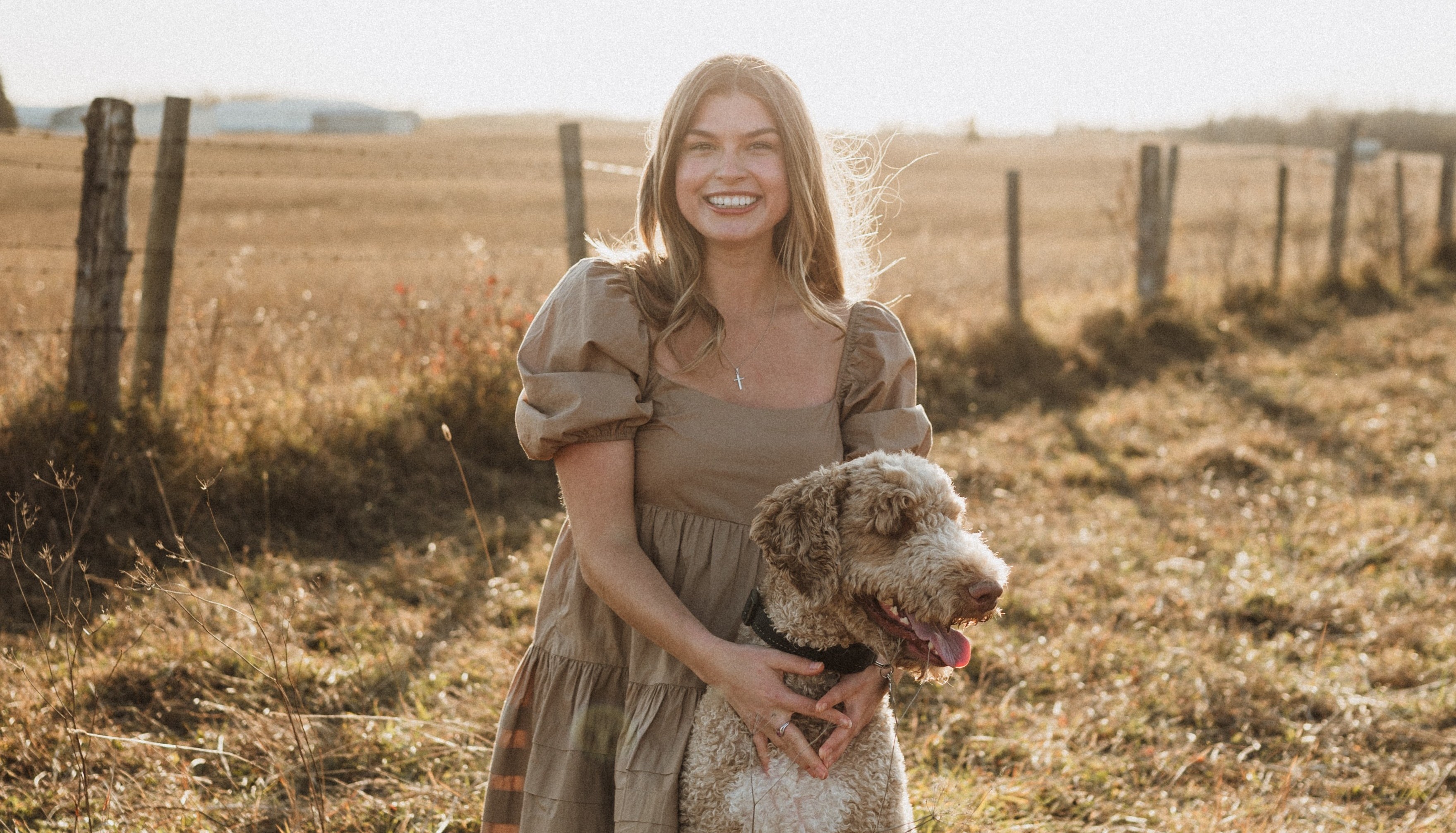 Hope Grootenboer, in a field during the autumn, puts her arms around a dog; a wire fence is behind her