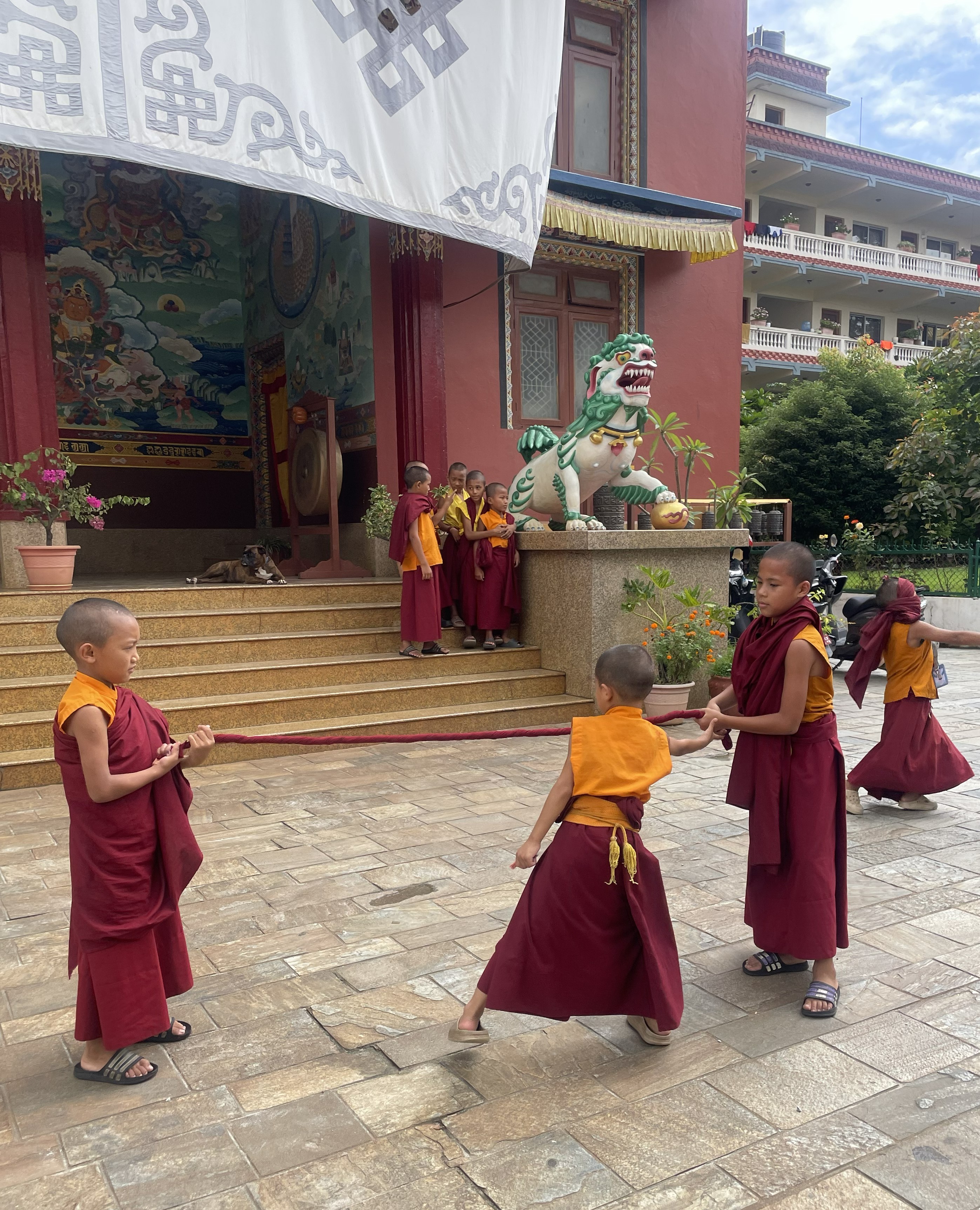 Small boys wearing red and yellow robes stand on the steps of a building while another group of boys plays on the building's forecourt
