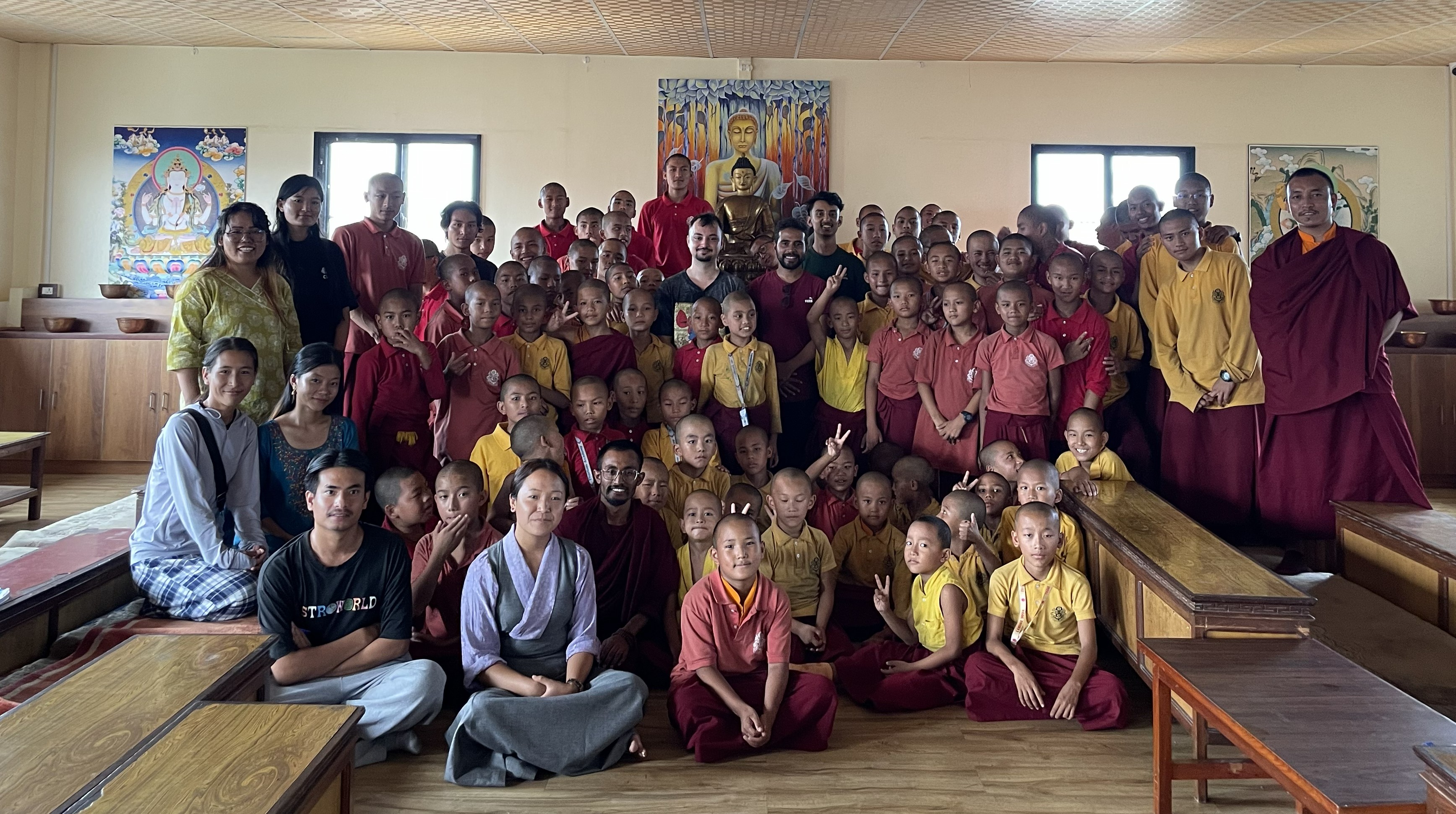 Students and members of the Tharlam Monastery pose for a group photo in a room with images of Buddha on the wall