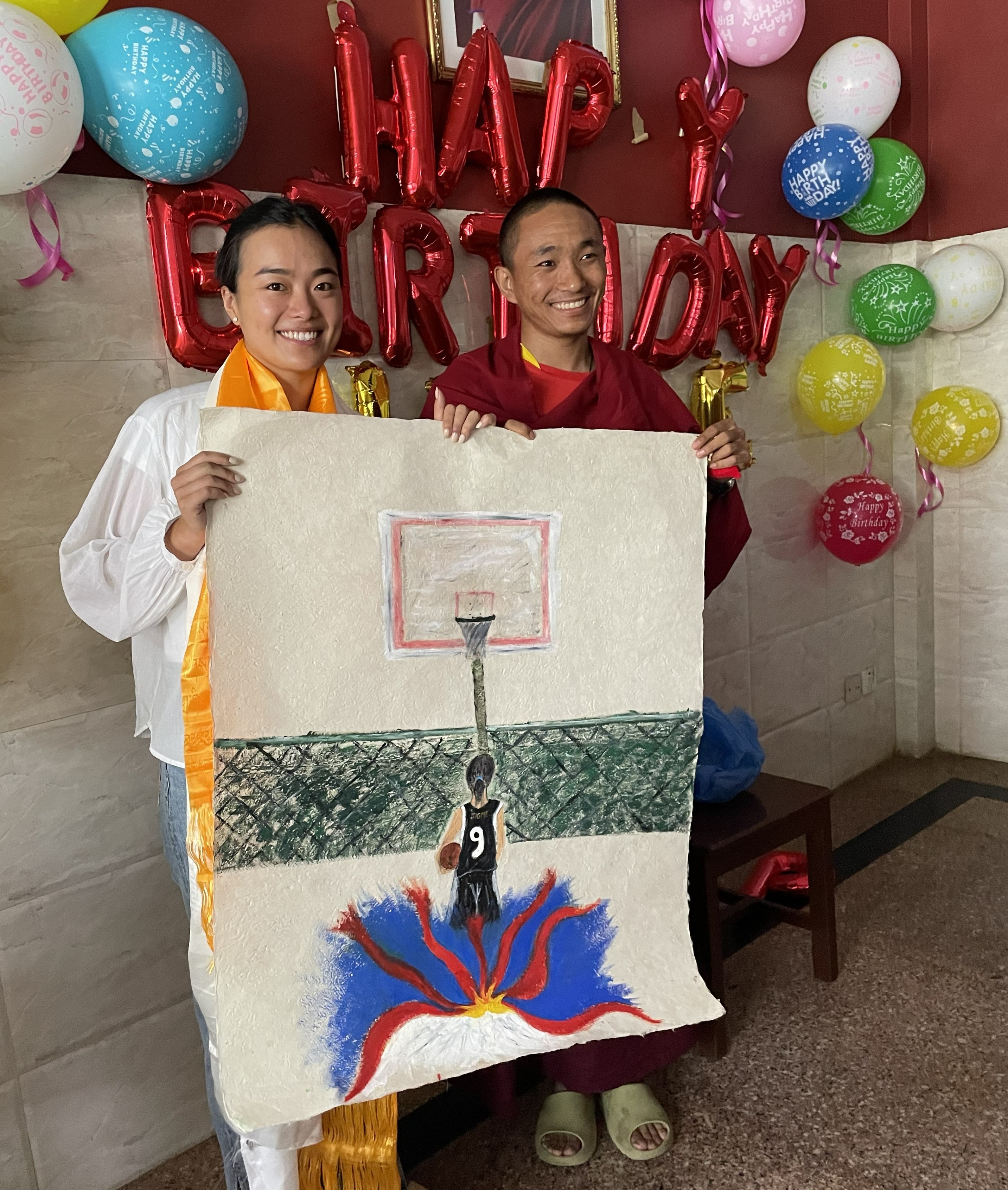 Jigme Tsering and a Buddhist monk stand in a room with balloons saying Happy Birthday and hold up a drawing of a basketball player