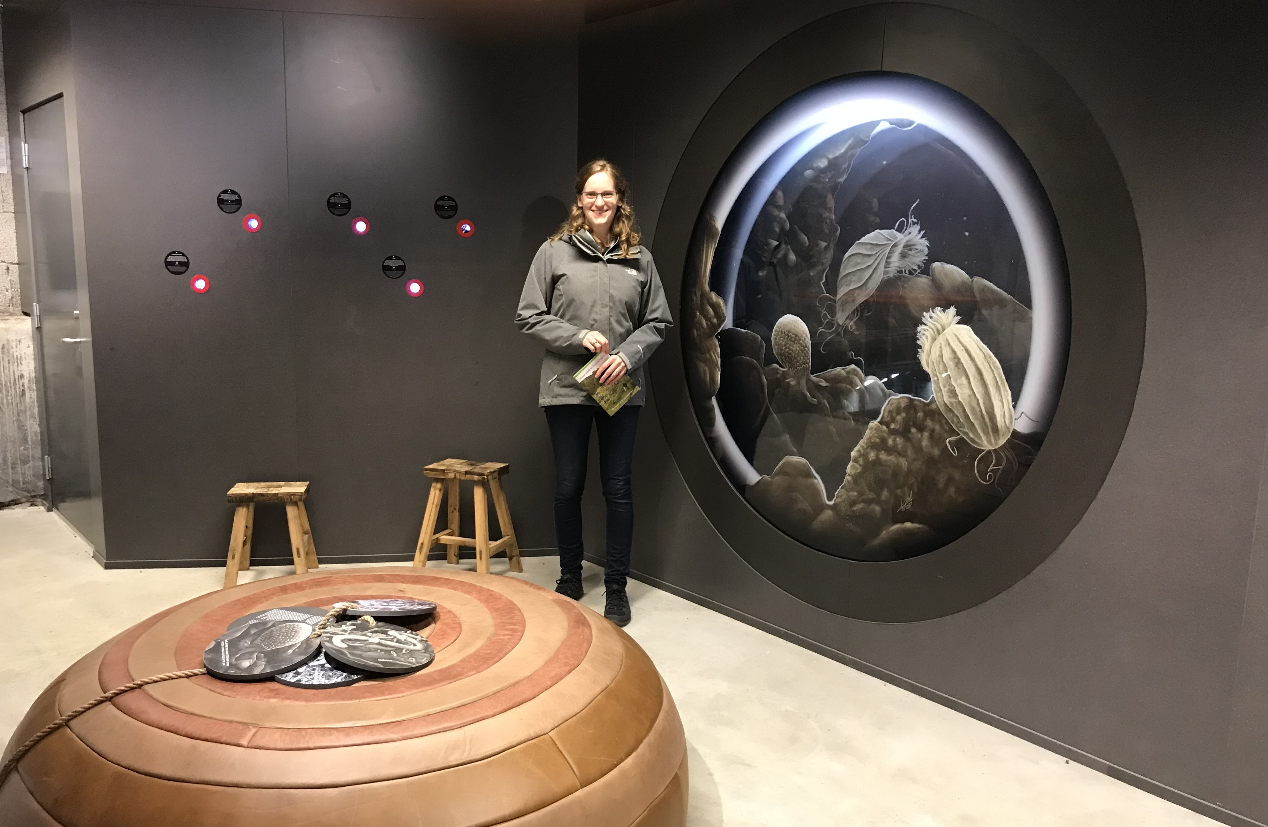 Kate Solbakk at Art Exhibit Katte Solbakk stands beside a very large wall-mounted circular illustration of protozoa; in front of her are smaller round protozoa images attached to each other by a cord sitting on a round cushioned object