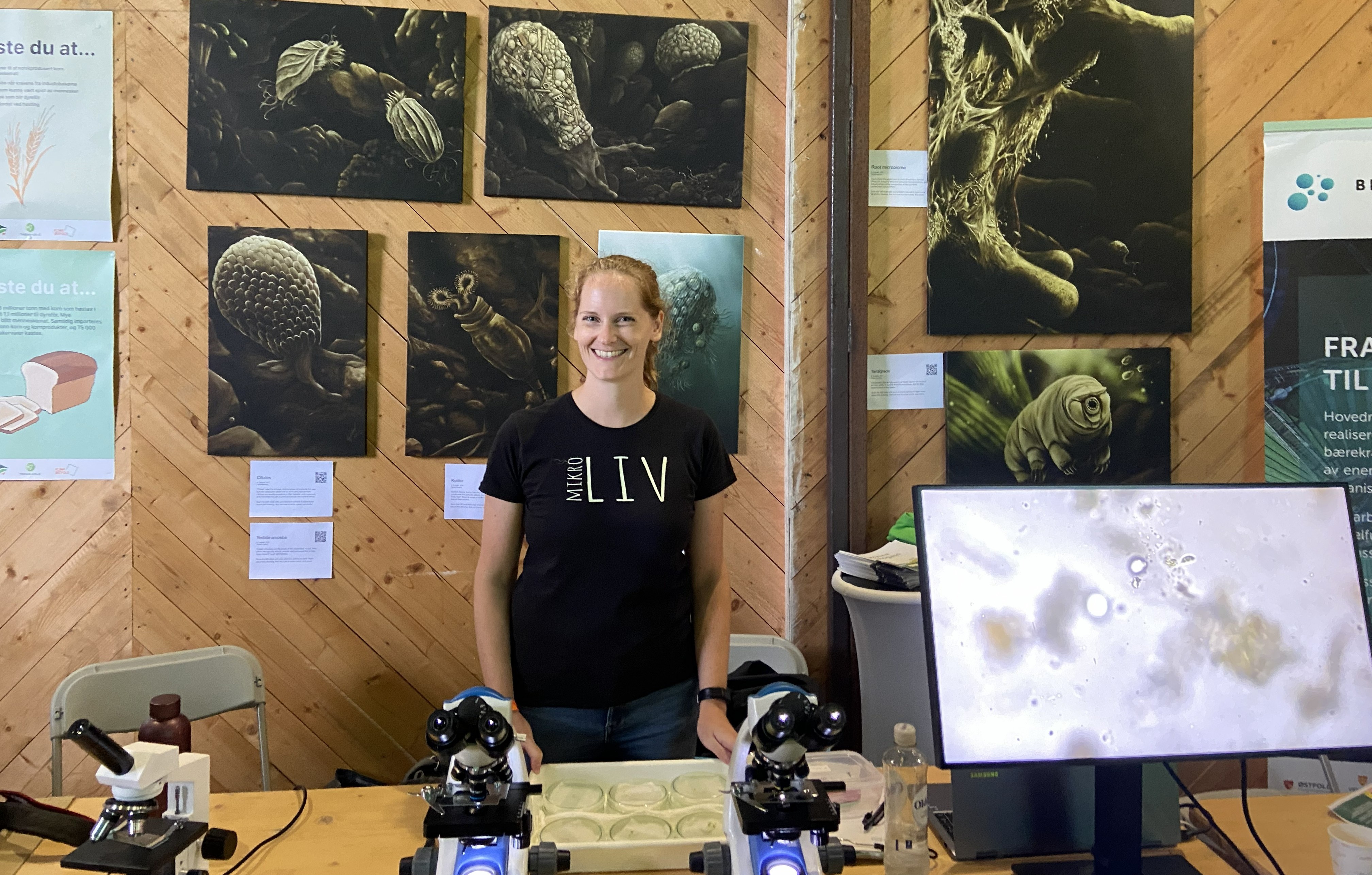 Kate Solbakk with Scientific Display Kate Solbakk, wearing a black t-shirt with the words "Mikro Liv" on it stands behind a table with microscopes; microscopy illustrations hang on the wall behind her