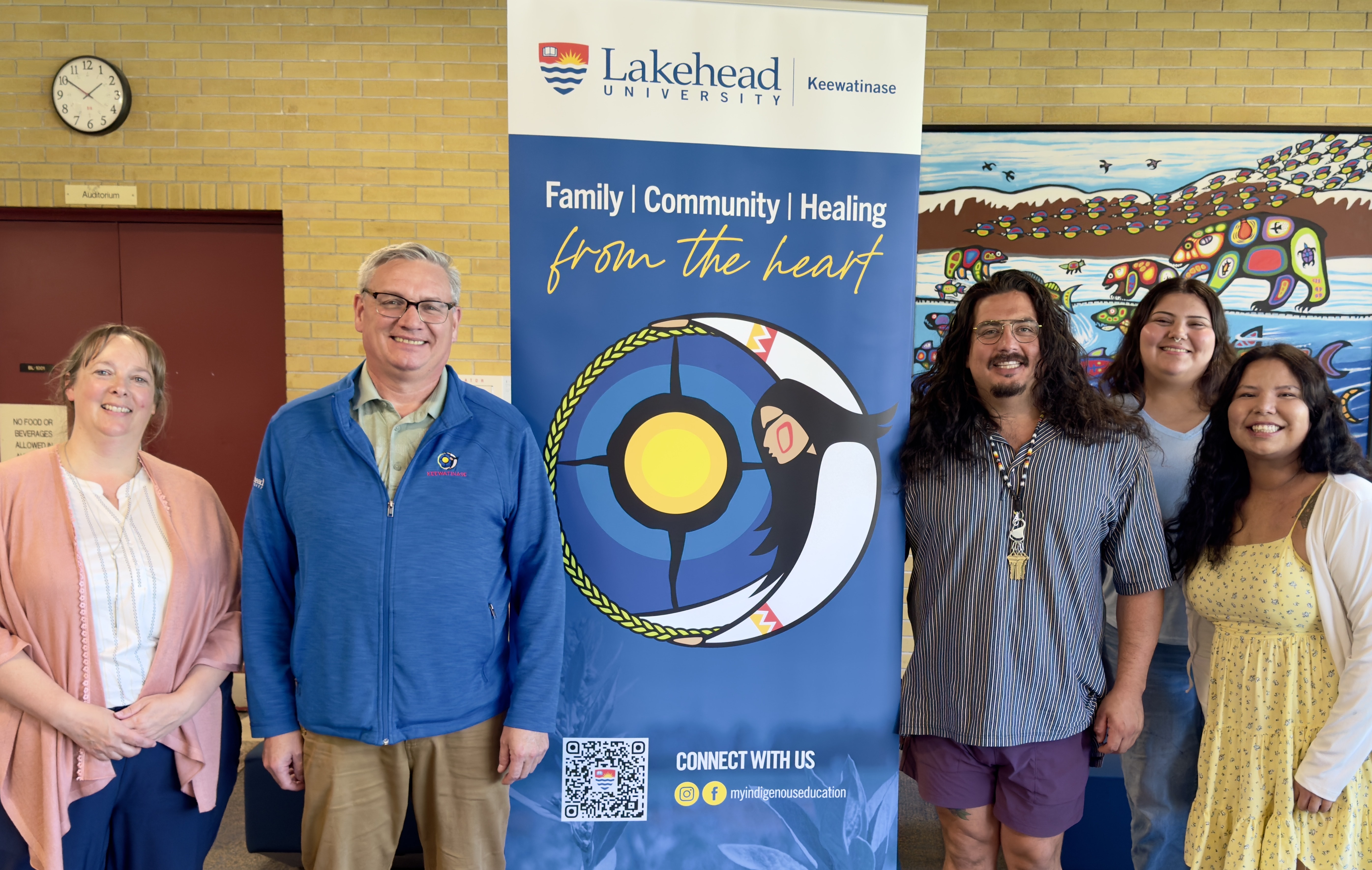 Keewatinase Team Melissa Oskineegish, Dr. Paul Cormier, Tyler Armstrong, Nevaeh Rae, and Roberta Ogemah stand in front of the Bora Laskin Building auditorium beside a vertical banner with information about the Keewatinase program