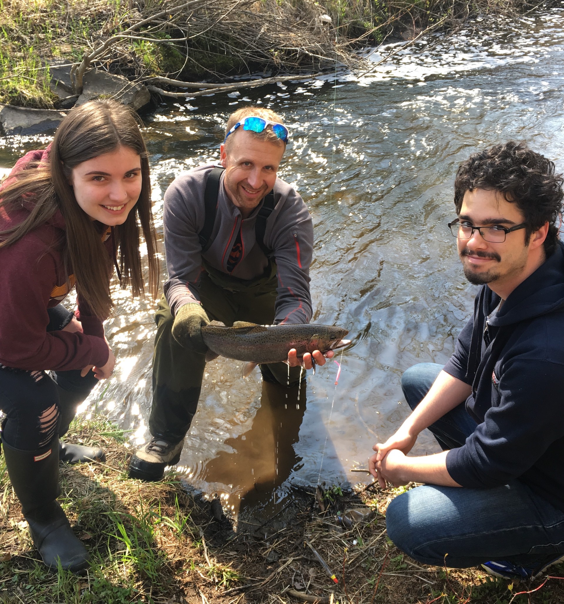 Keith Ailey Fishing with Students Keith Ailey crouches in a stream while holding a speckled fish in his hands; a student stands on either side of him