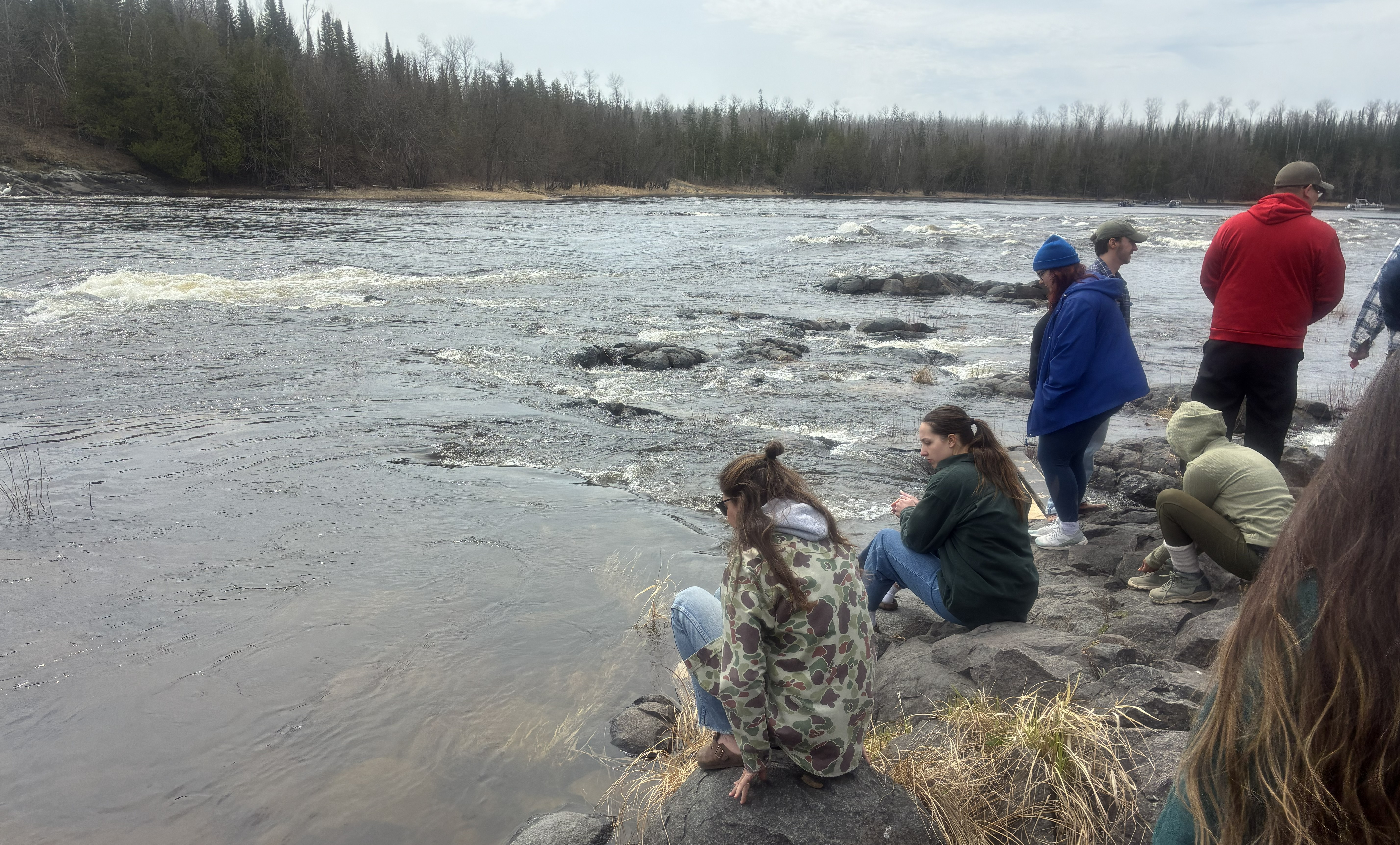 A back view of a group of people sitting and standing on rocks at the edge of a river