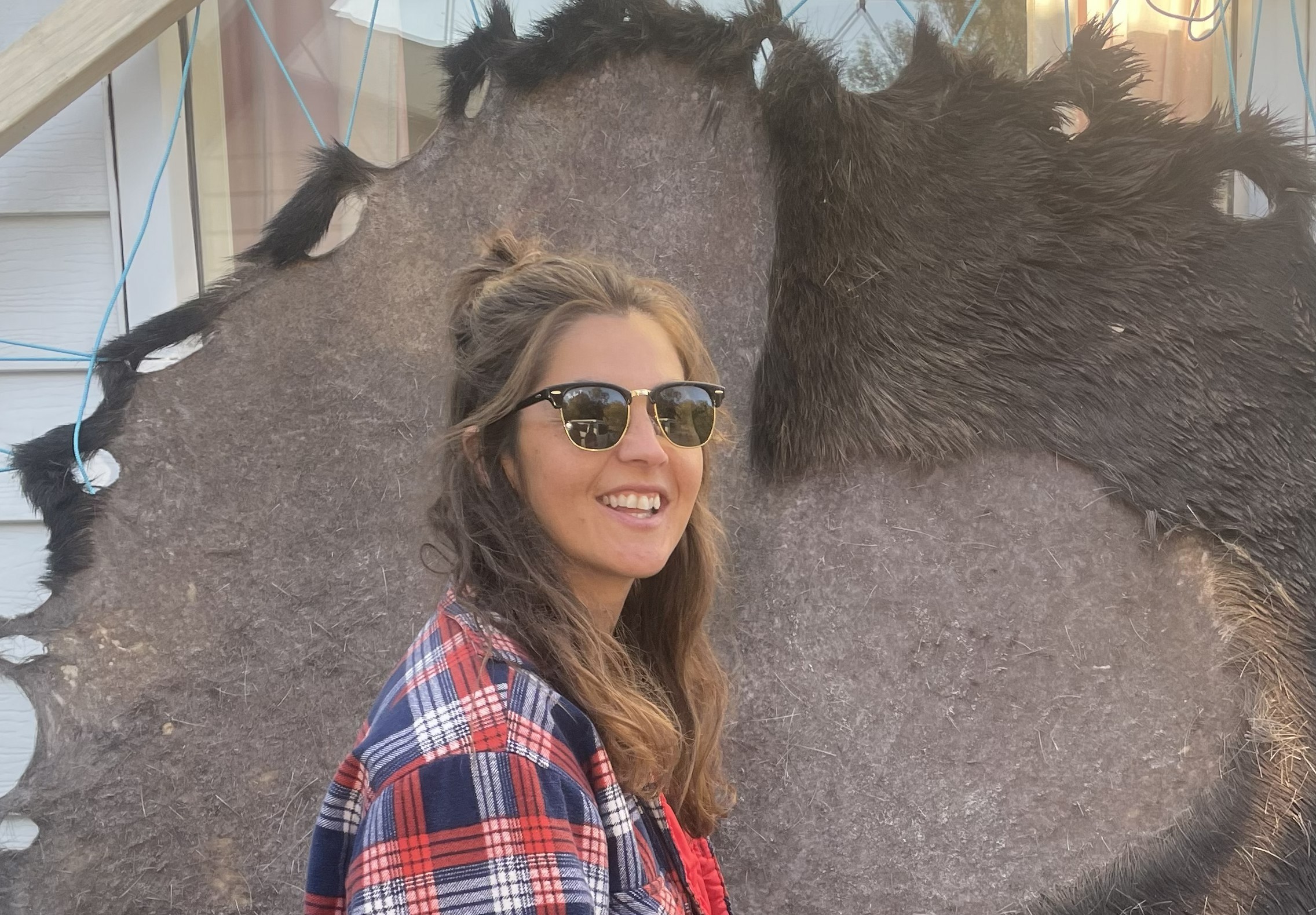 Larissa Speak, wearing a plaid jacket and sunglasses, stands in front of a moose hide that is being tanned