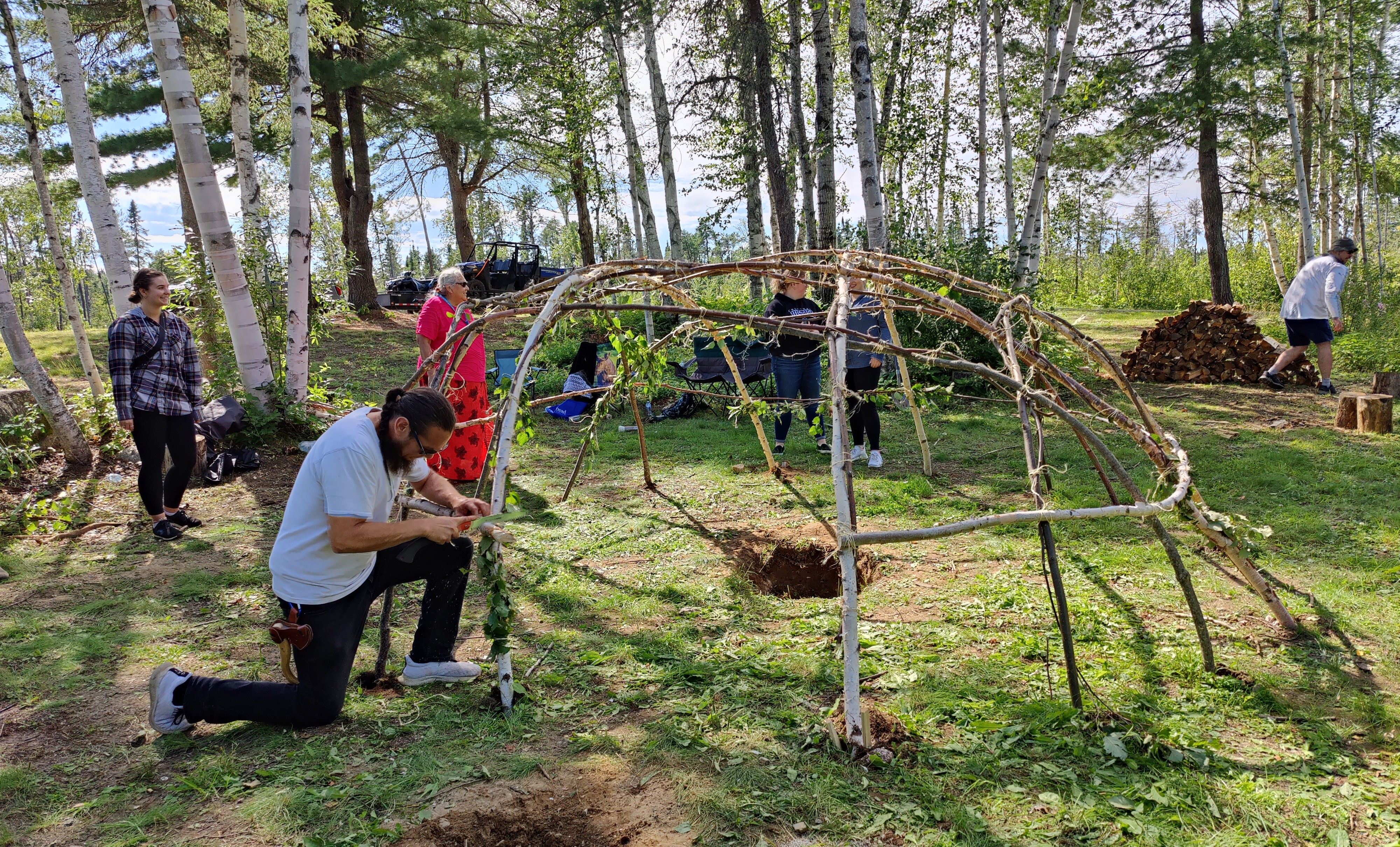 In a clearing surrounded by trees, Robin Sutherland constructs a sweat lodge out of tree saplings while several other people stand on the edge of the clearing