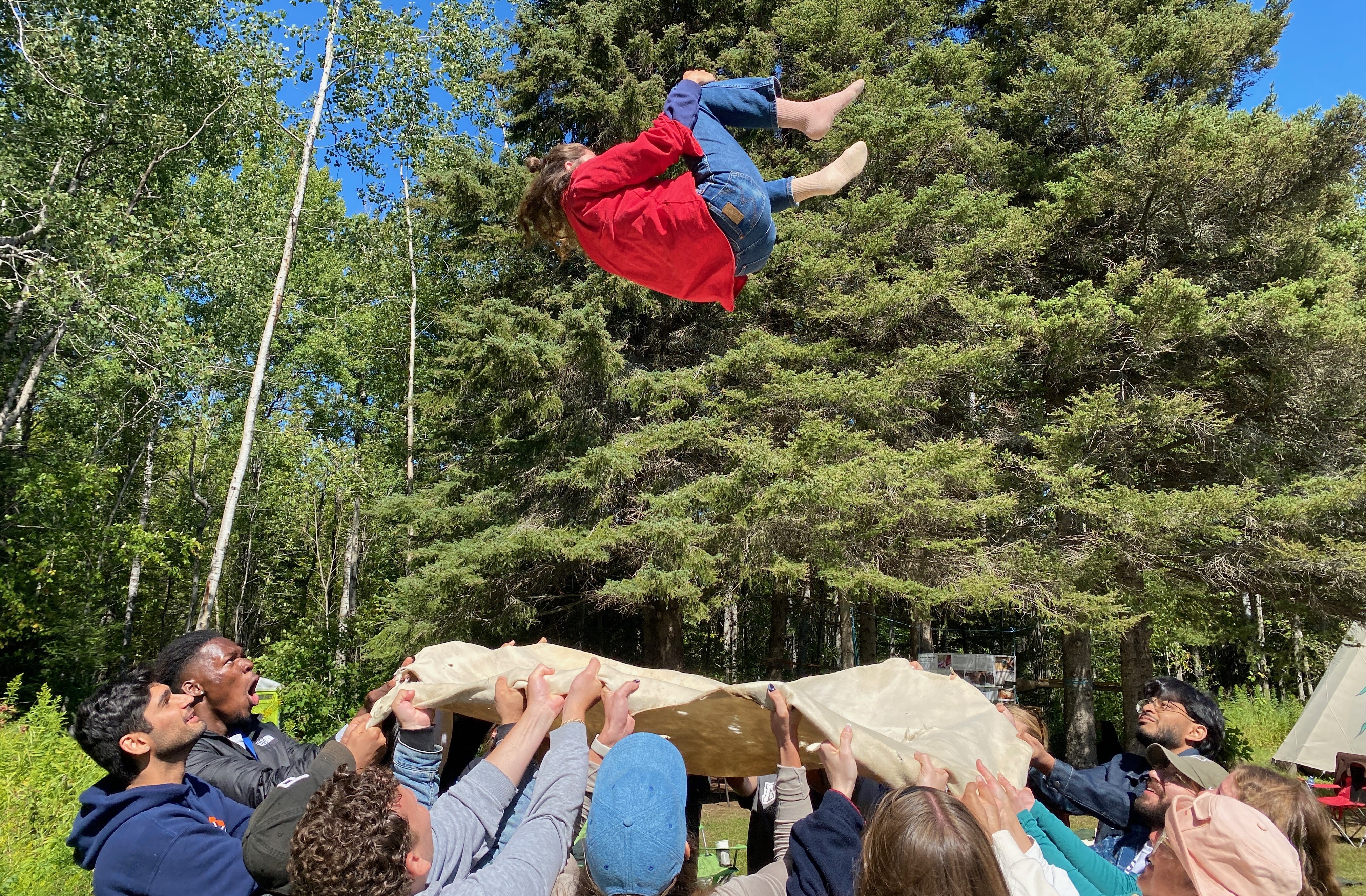 A group of people stand in a circle holding a moose hide and toss a woman from the moose hide up into the air