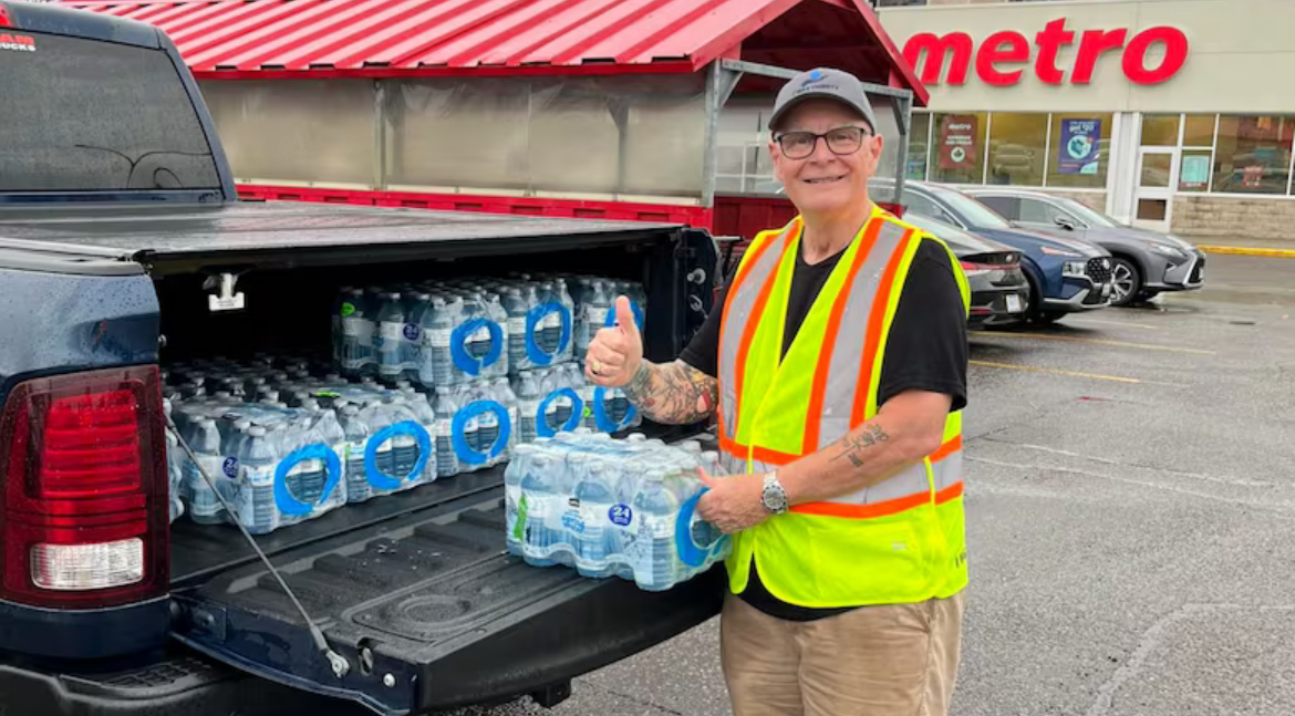 Lorne Clifford, wearing a safety vest and baseball cap, gives the thumbs up signal while standing beside the back of pickup truck filled with cases of bottled water in the parking lot of a Metro grocery story