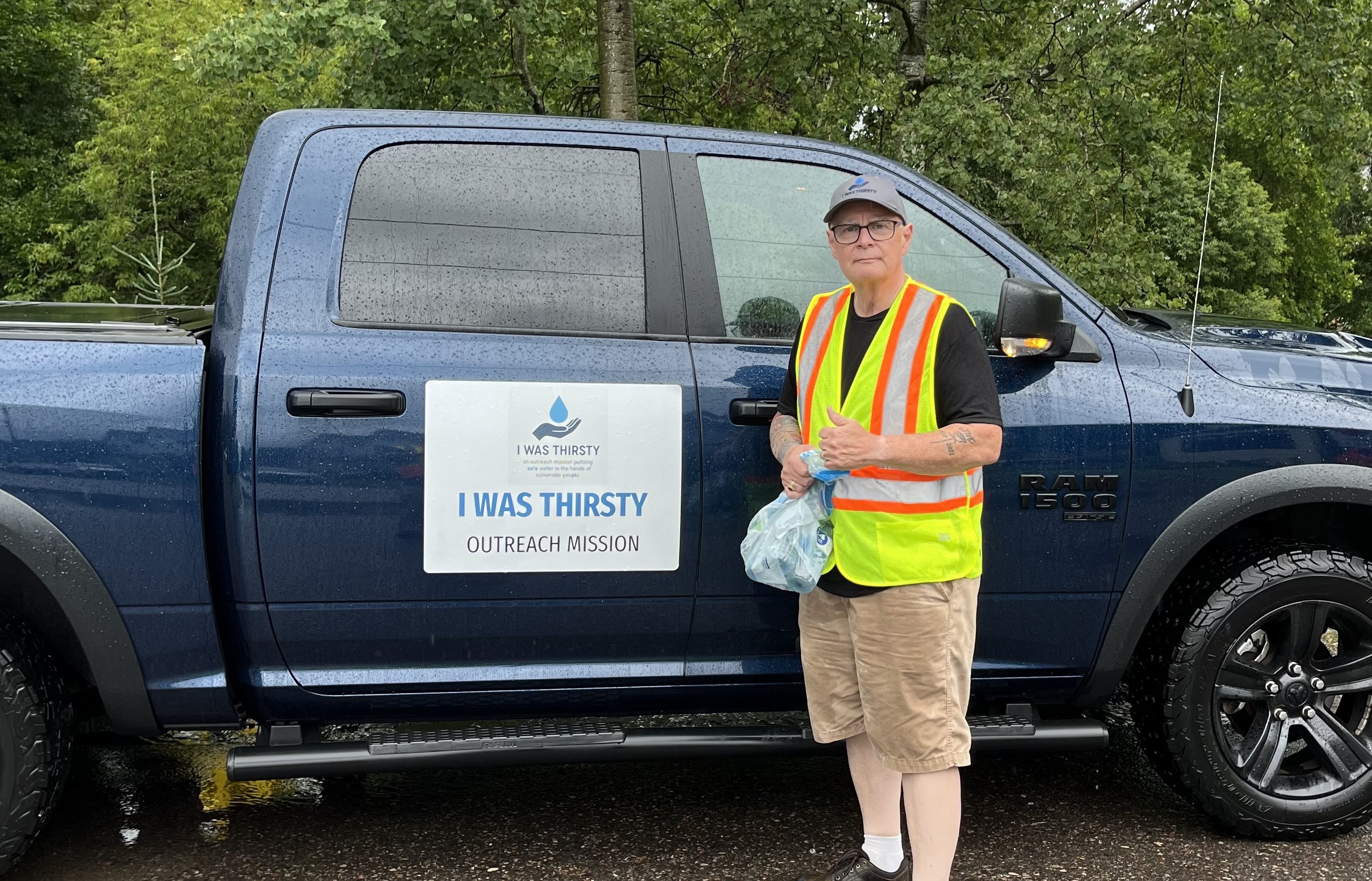 Lorne Clifford, wearing a safety vest, stands beside a dark blue RAM pickup truck with a logo saying I WAS THIRSTY OUTREACH MISSION