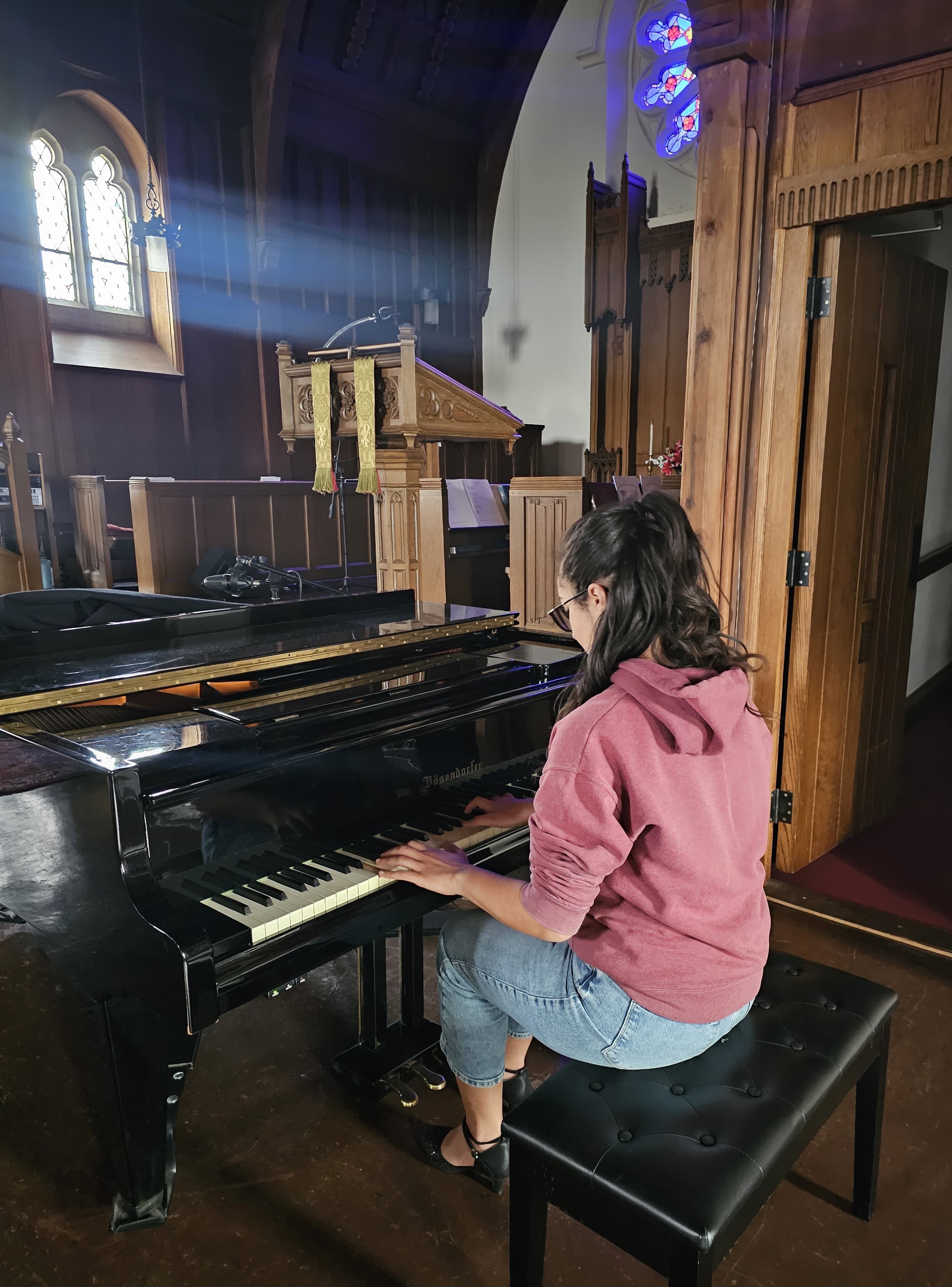 Mary Clark plays the piano in an empty church with a stained glass window and an ornate podium and wall mouldings