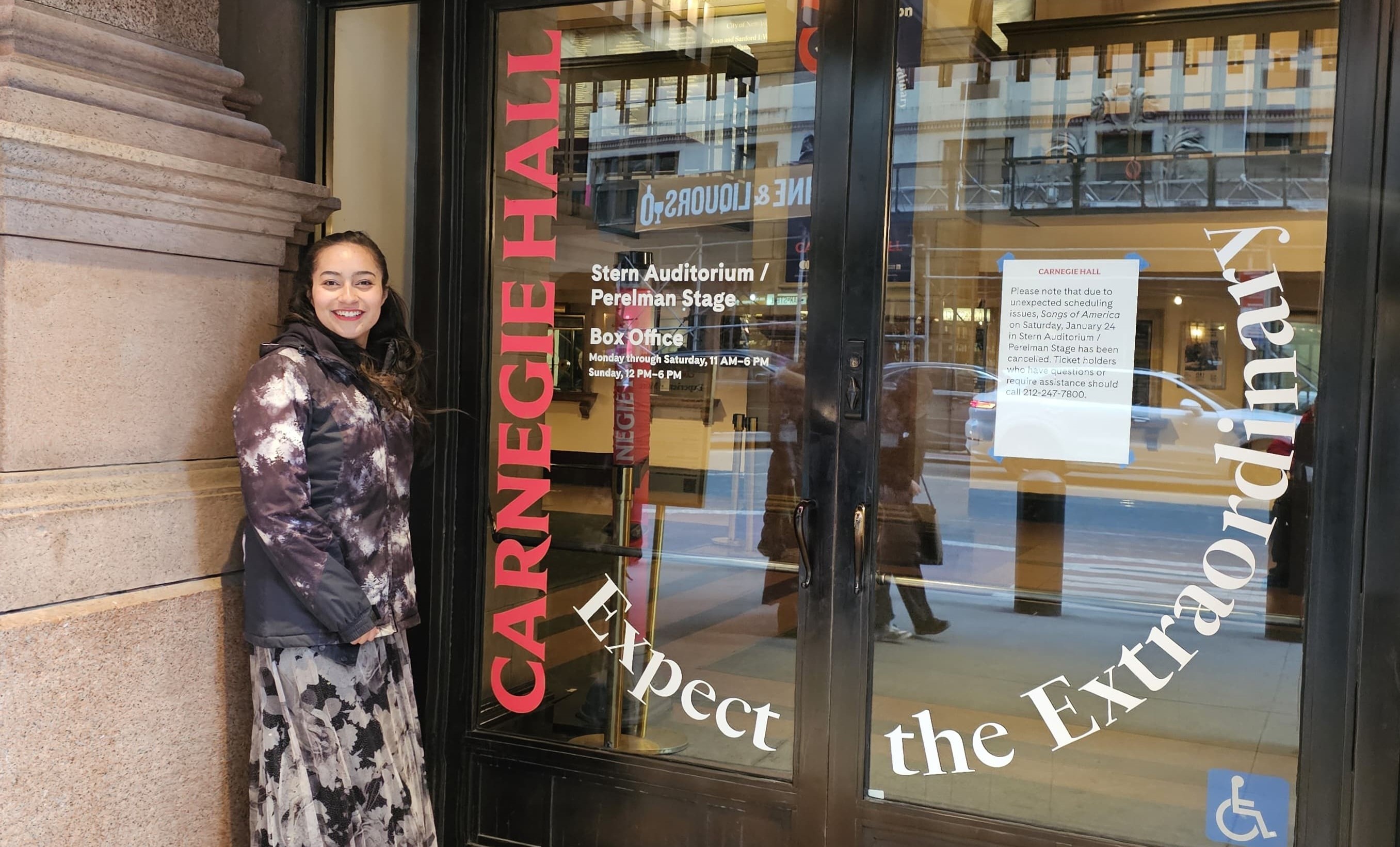 Mary Clark stands beside exterior glass doors emblazoned with the words Carnegie Hall Expect the Extraordinary