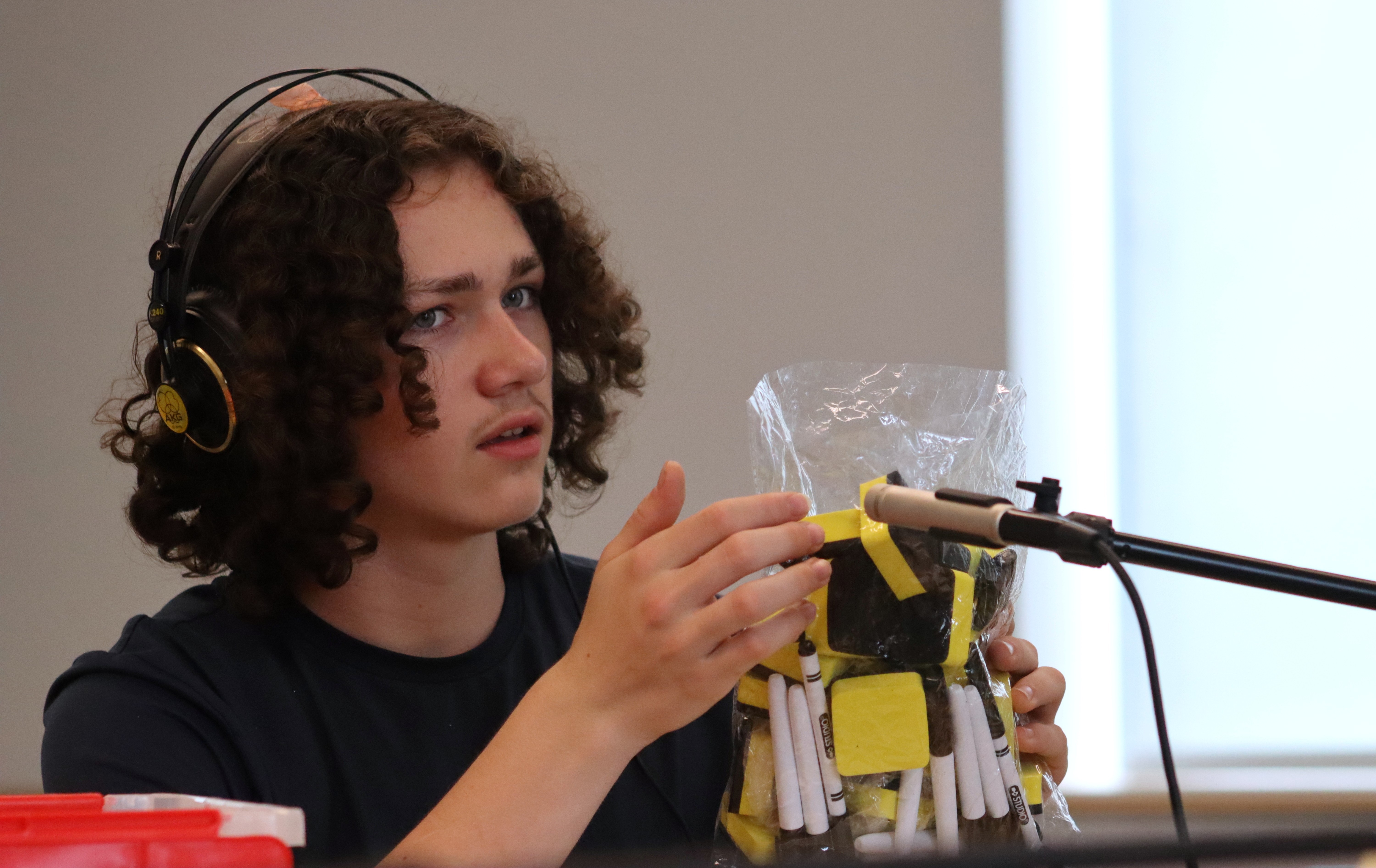 A teenage boy with brown curly hair wears headphones and holds a plastic bag filled with markers and other items in front of a microphone