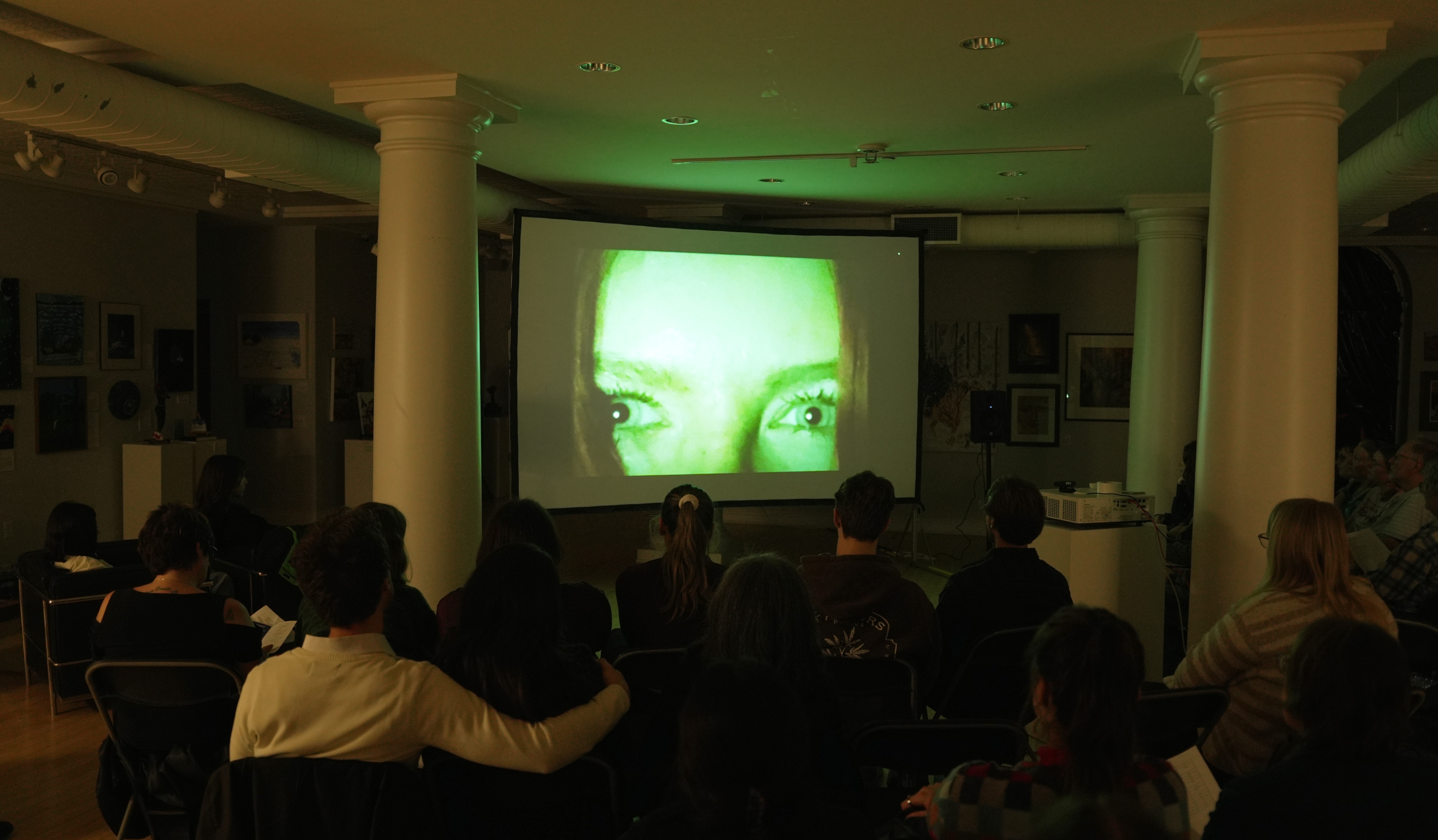 A back view of a group of seated people in a room with columns and artworks on the walls watch a movie screen with a close up of a woman's face