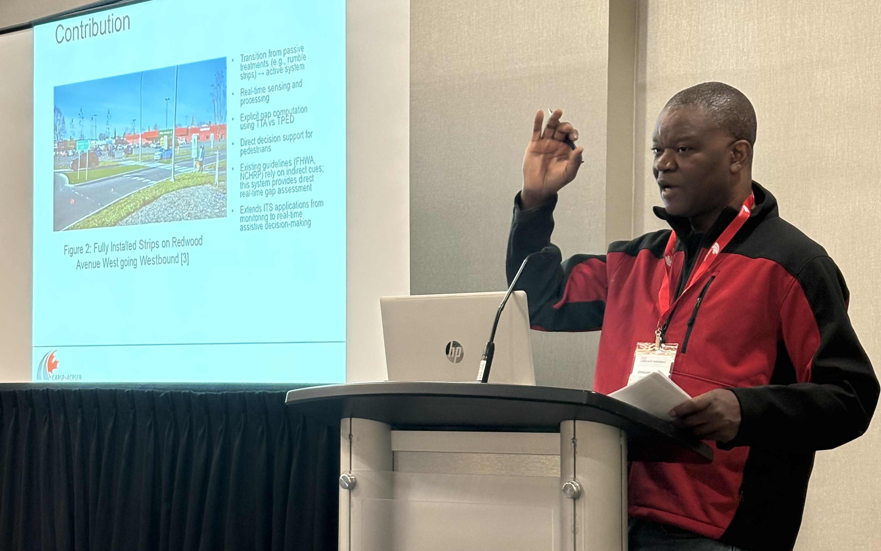 Omotunde Adeniran Gives CARSP Presentation Omotunde Adeniran, wearing a red and black fleece jacket, speaks at a podium; a screen displaying a presentation slide is next to the podium