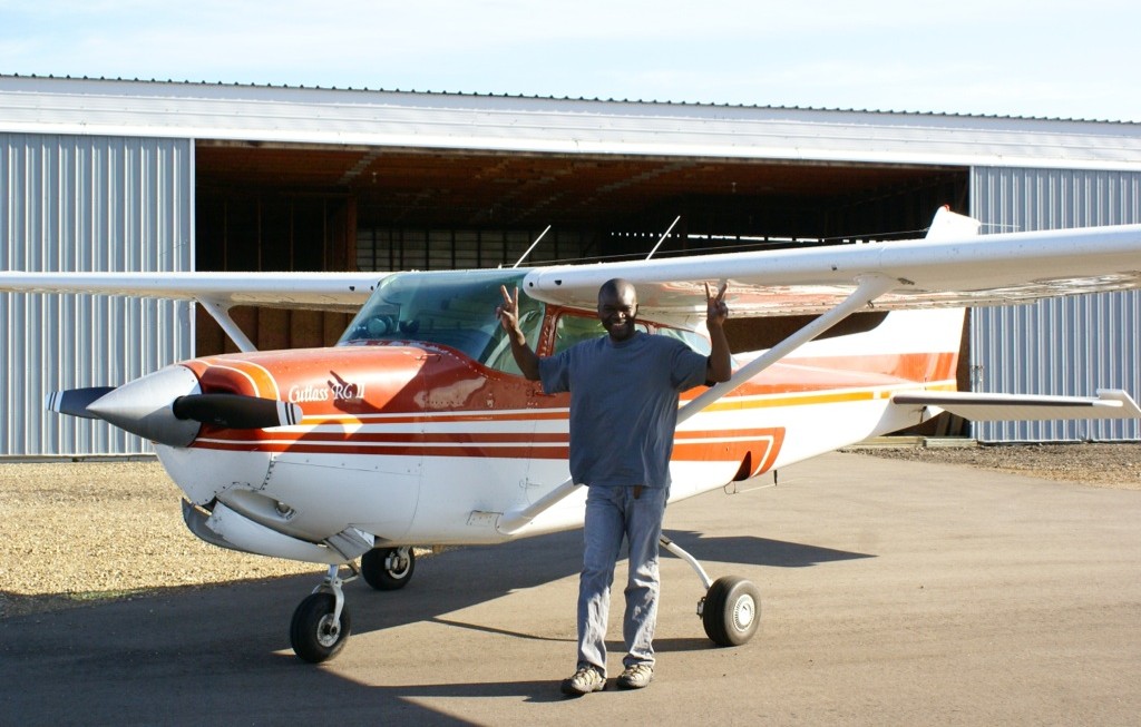 Omotunde Adeniran with Plane Omotunde Adeniran stands in front of a small plane with both hands raised in the victory sign; behind the plane is a metal hangar