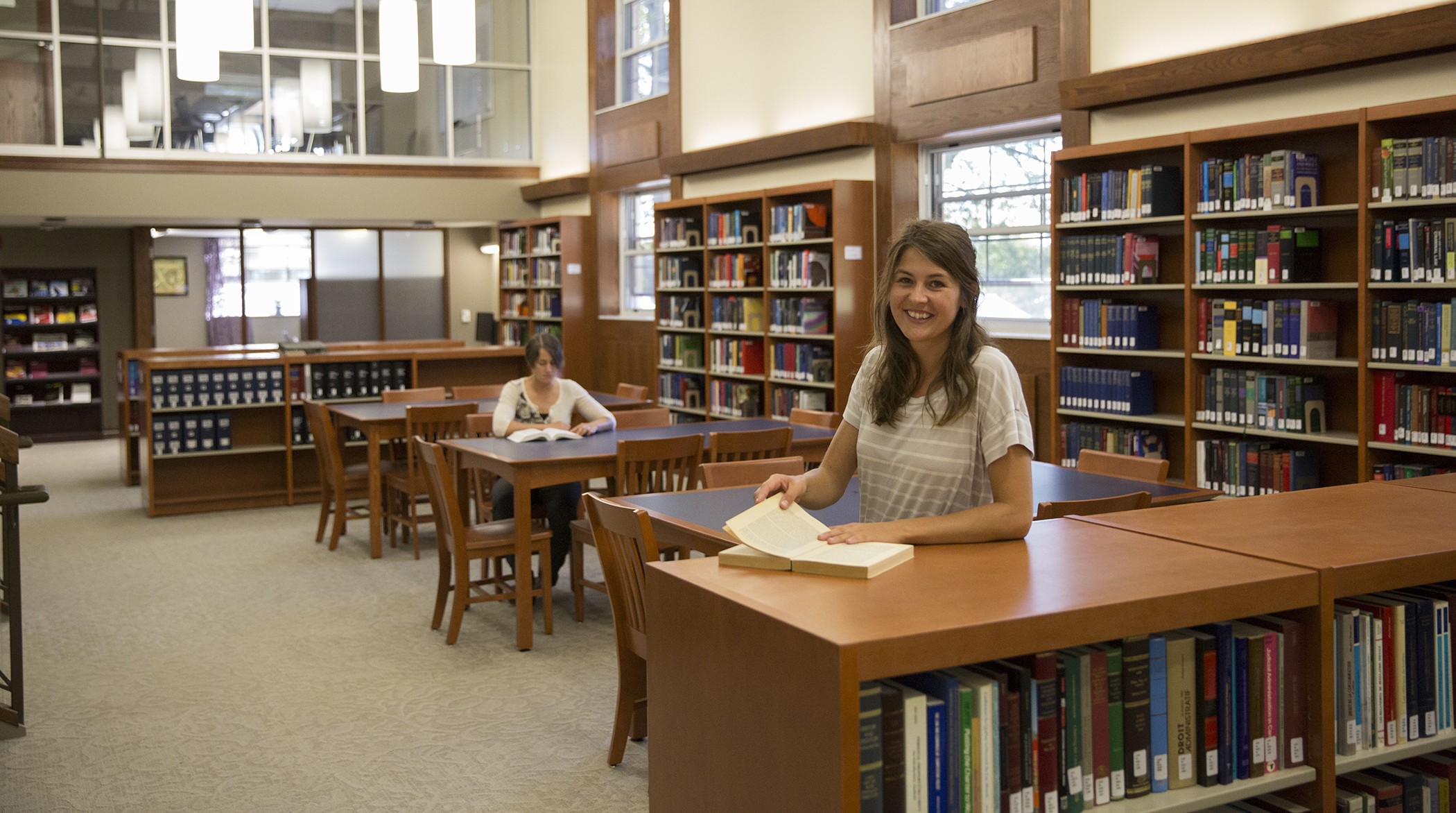 An interior view of the Bora Laskin Faculty of Law Library with a young woman standing behind a low bookcase with an open book and a seated woman reading a book at a table in the background