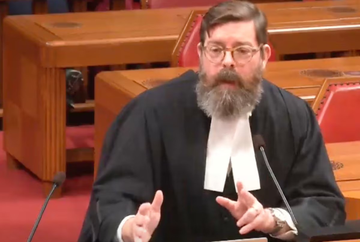 Ryan Alford, wearing glasses, a black robe, and a white jabot, sits a table with two microphones in a red-carpeted courtroom with empty wooden tables and chairs behind him