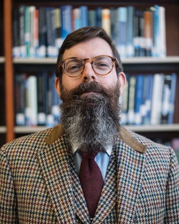 Ryan Alford, wearing glasses, a tie, and a houndstooth-checked vest and blazer, stands in front of a bookcase