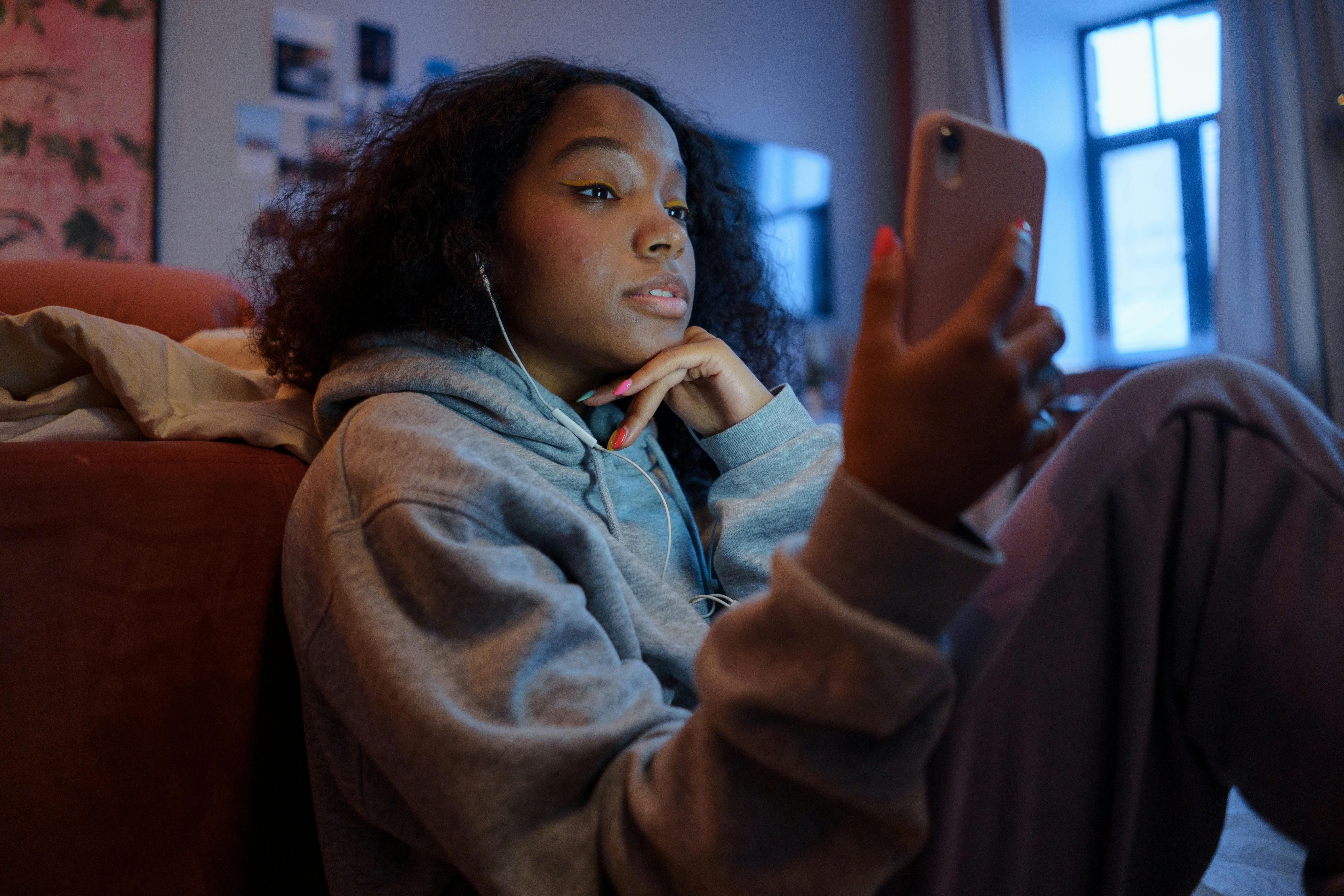 A teenage girl wearing headphones sits on the floor in a darkened room looking at an iPhone in her hand