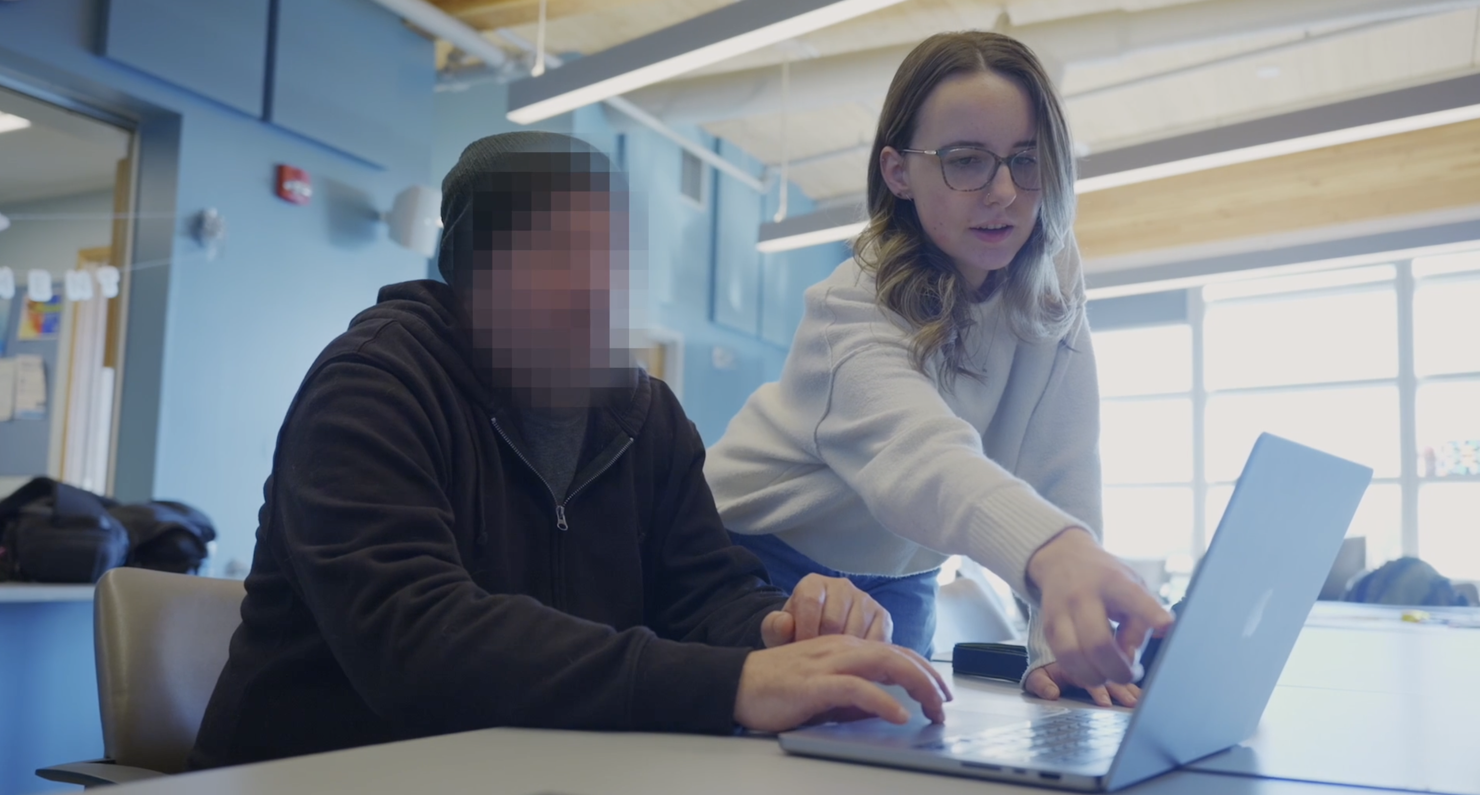 Sydney Ambury with Digital Storyteller Sydney Ambury, wearing a white sweater and glasses, points at a laptop screen while standing beside a man wearing a black hoodie and a black hat who is seated in front of the laptop