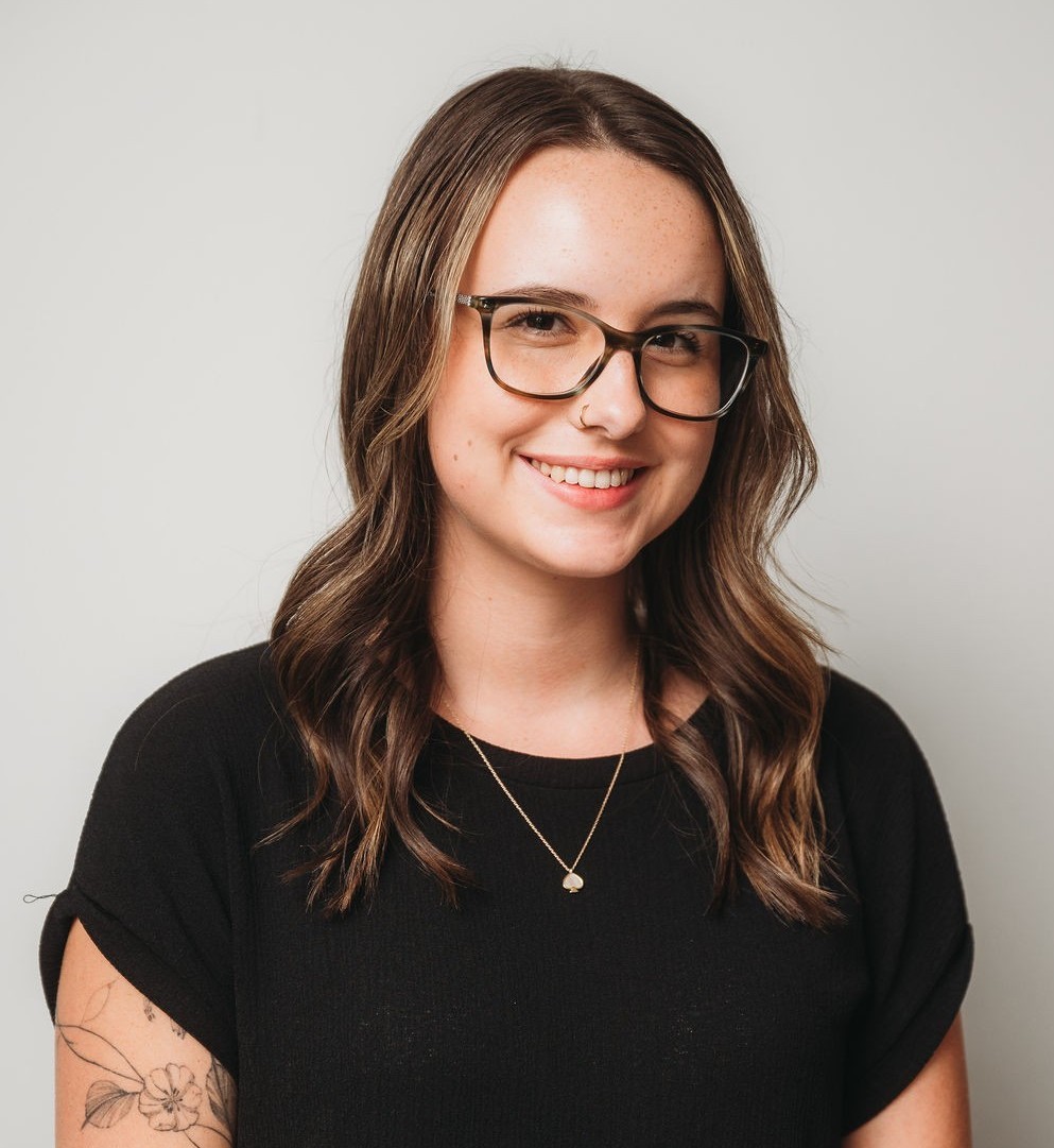 Sydney Ambury A head-and-shoulders photo of Sydney Ambury smiling and wearing a black shirt, a pendant, and glasses