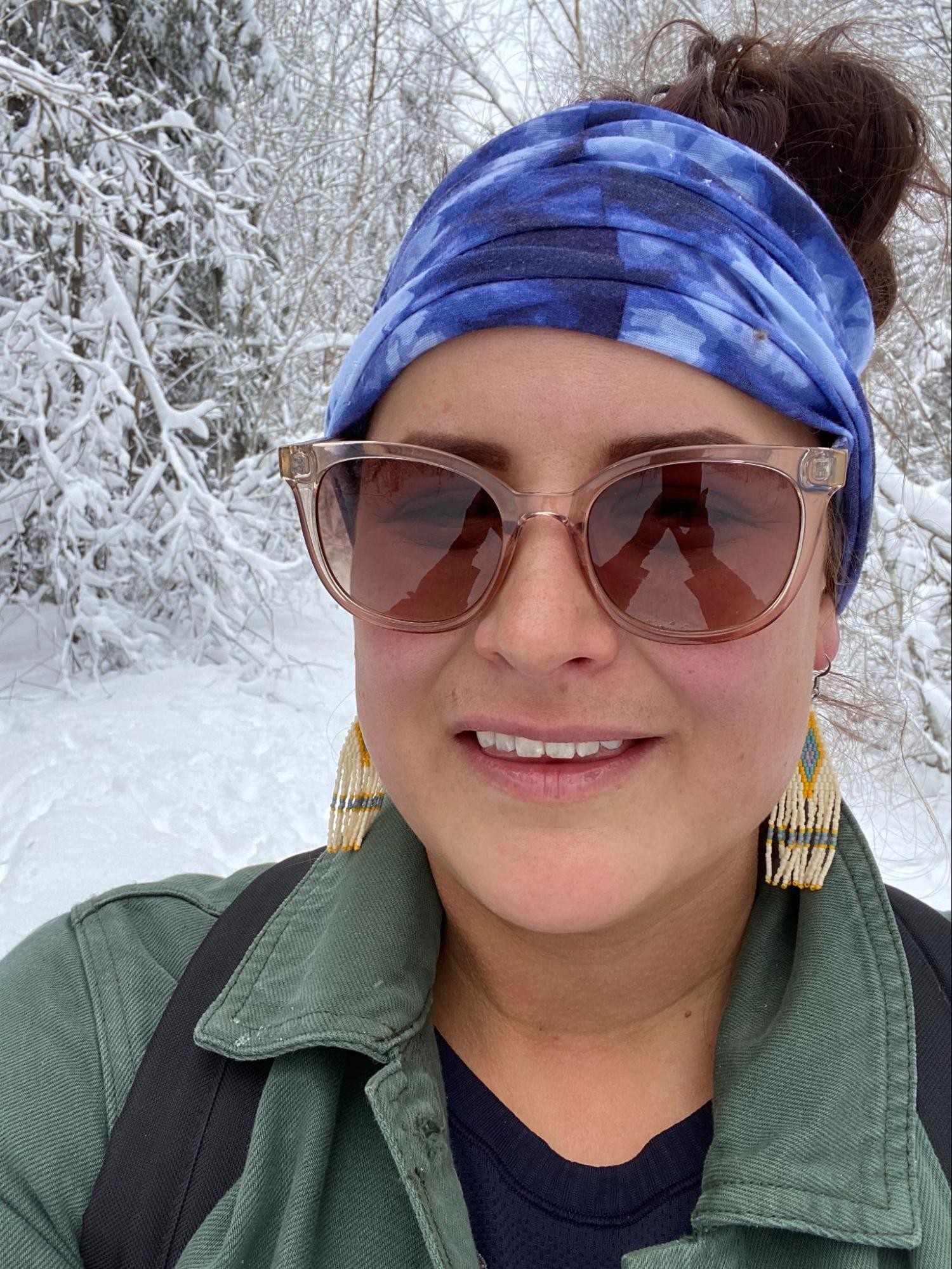 A head-and-shoulders photo of Tamara Bernard, wearing sunglasses and beaded earrings, standing in front of snow-covered trees