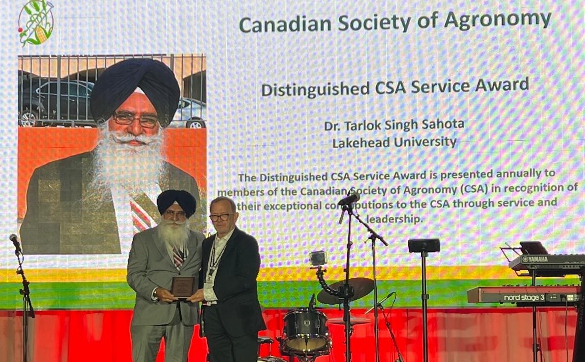 Dr. Tarlok Singh Sahota stands next to another man on a stage while holding an award plaque; behind them is a large screen with Dr. Singh Sahota's photo and text saying that Dr. Singh Sahota is the winner of the Distinguished CSA Service Award