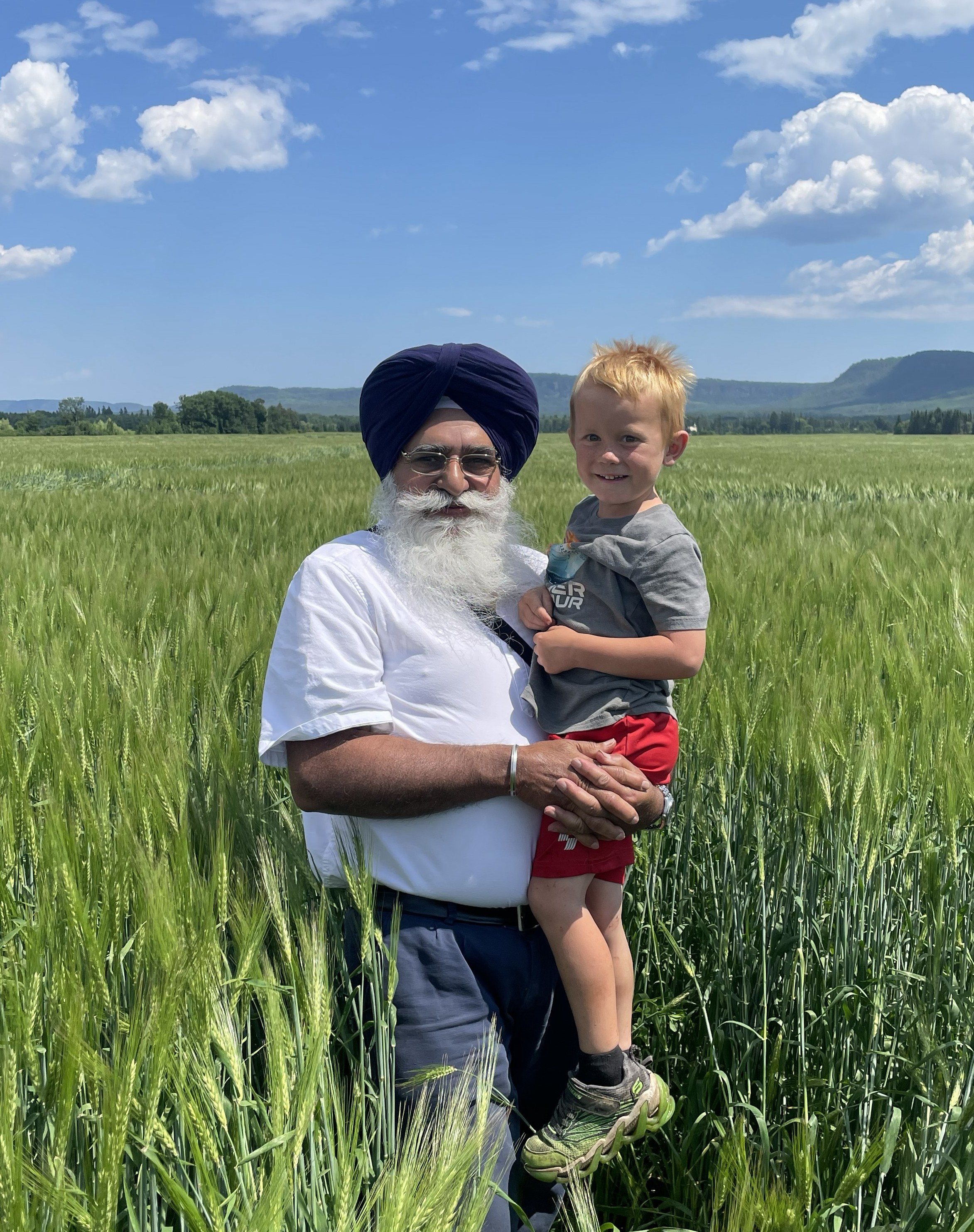 Dr. Tarlok Singh Sahota stands in a barley field on a sunny summer day and holds a small smiling boy in his arms
