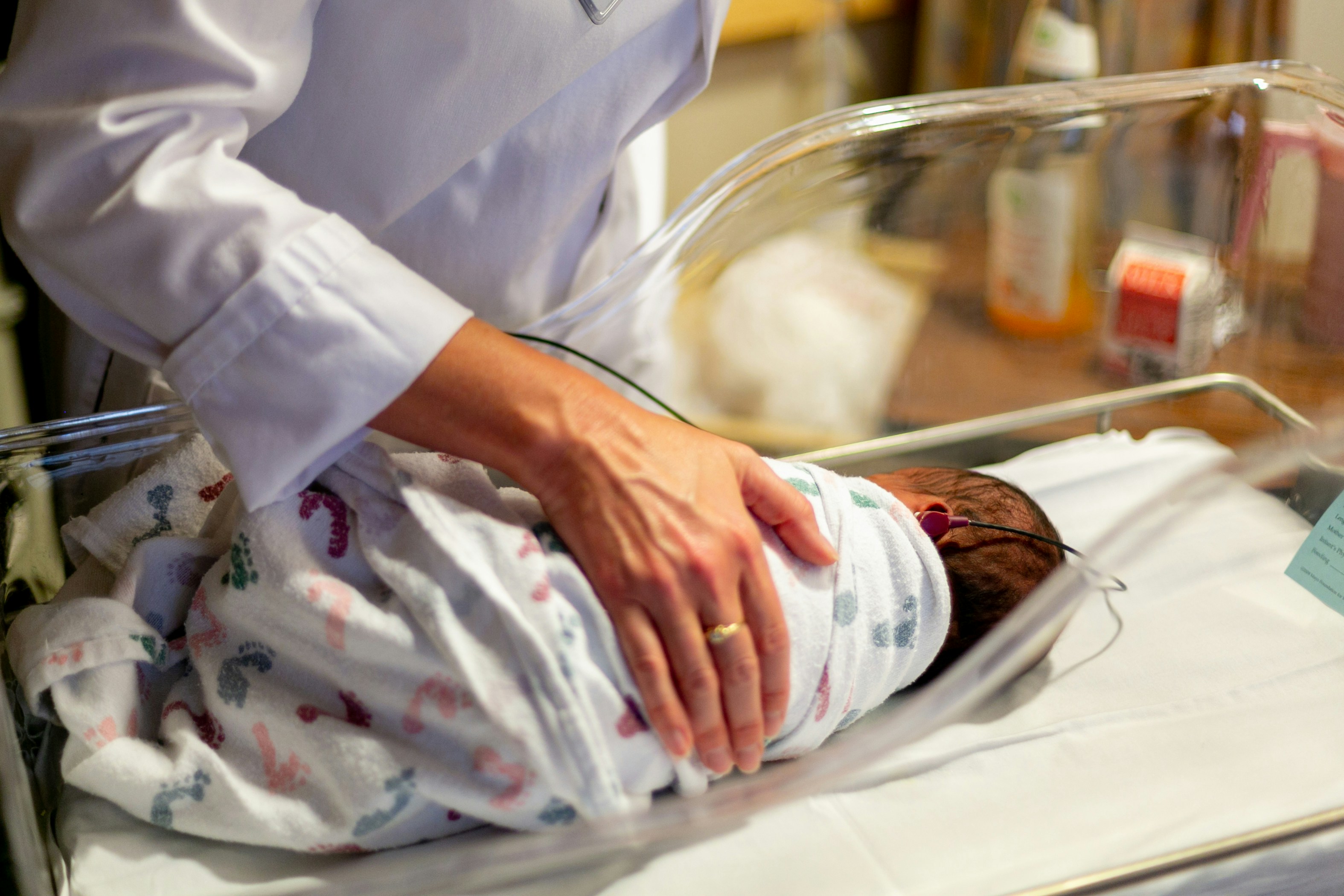 Baby in Incubator A woman wearing a white medical coat stands over an incubator with her hand on an infant swaddled in a blanket