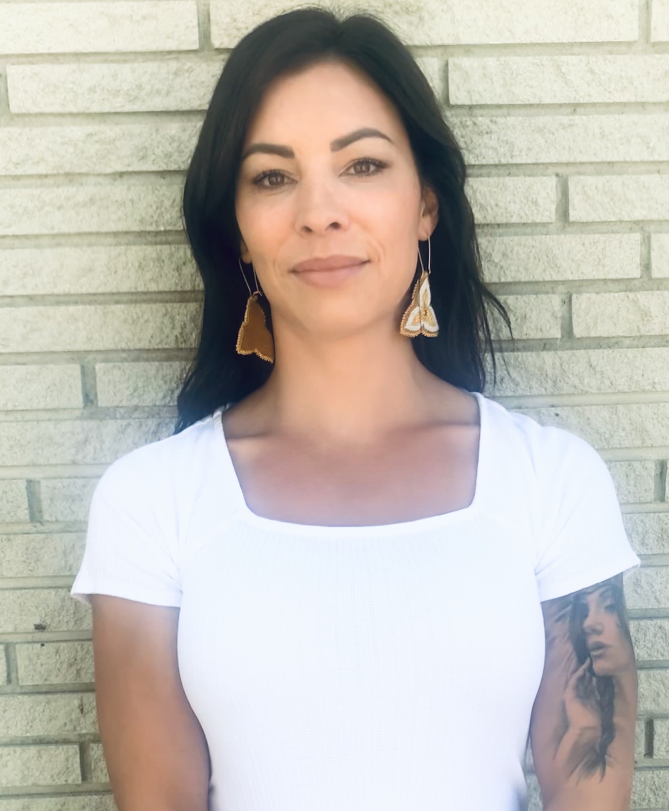 A head-and-shoulders photo of Dr. Zoe Michano-Furlotte, wearing a white t-shirt and beaded earrings, standing in front of a white brick wall