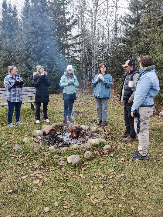 Indigenous Curriculum Specialist Joe Duncan (in baseball cap) shares teachings around a campfire, while participants drink cedar tea. 
